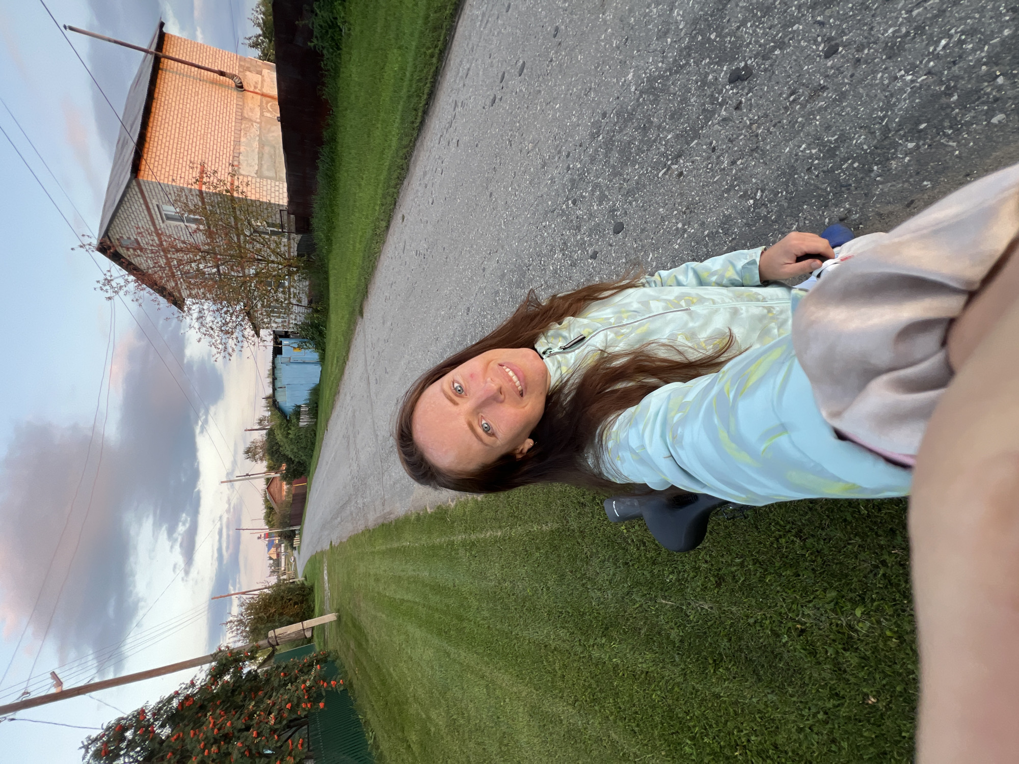 A woman is taking a selfie while sitting on a bicycle. She has long brown hair and is wearing a light blue jacket. The woman is smiling at the camera. The background shows a rural road in Pivkino, Russia, with houses and trees on either side. The sky is cloudy with hints of pink and orange from the setting sun. The image is taken from a low angle, looking up at the woman and the sky.
