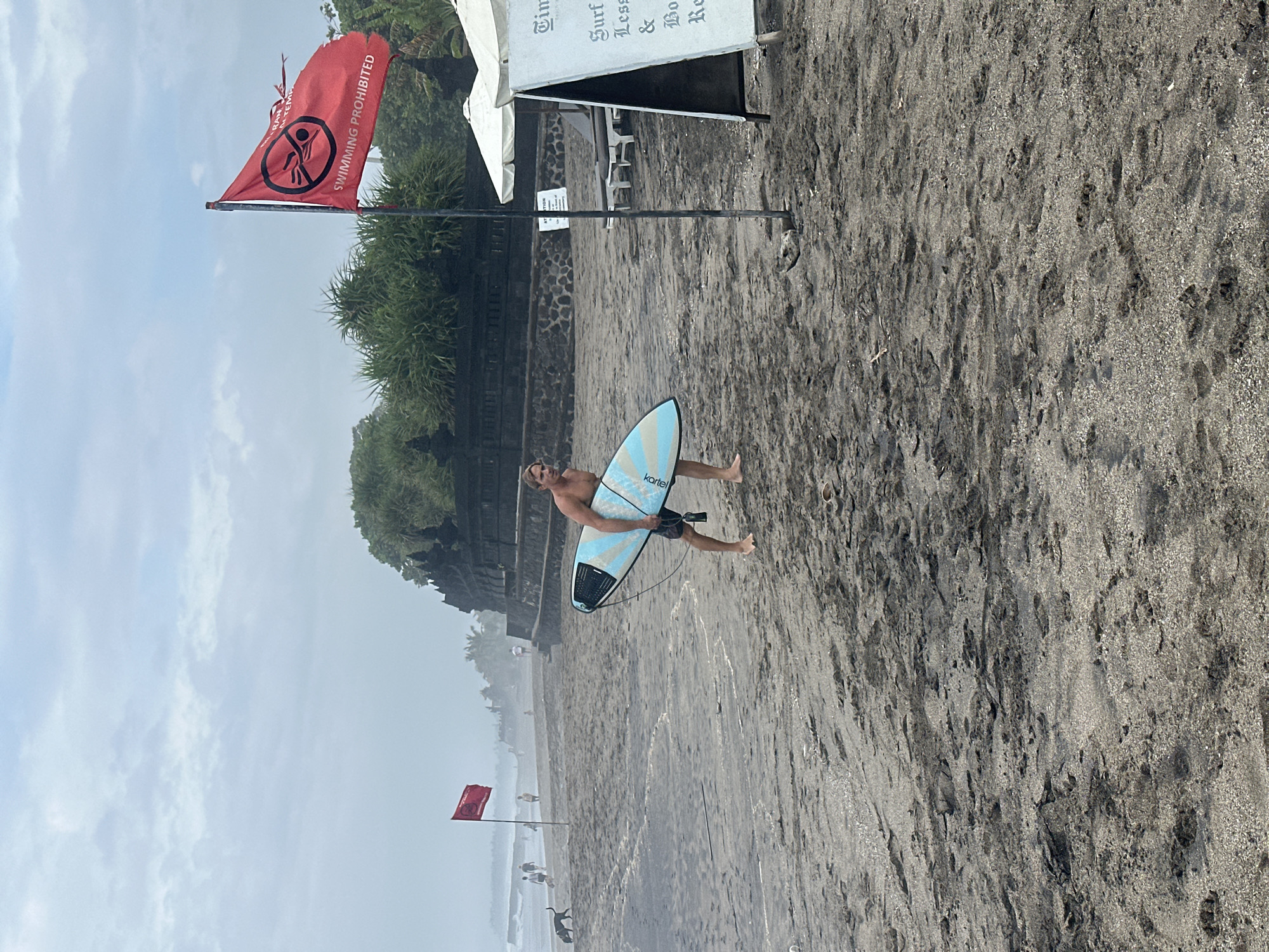 A man carrying a surfboard walks on a dark sandy beach near the water. He is wearing dark shorts and is barefoot. A red flag with a "swimming prohibited" symbol is visible in the background, along with lush green foliage and a stone structure that appears to be a temple. In the distance, other people and a dog are present on the beach. A sign with information about surf lessons and boat rentals is also visible. The sky is overcast, indicating a cloudy day. The scene is located in North Kuta, Indonesia.