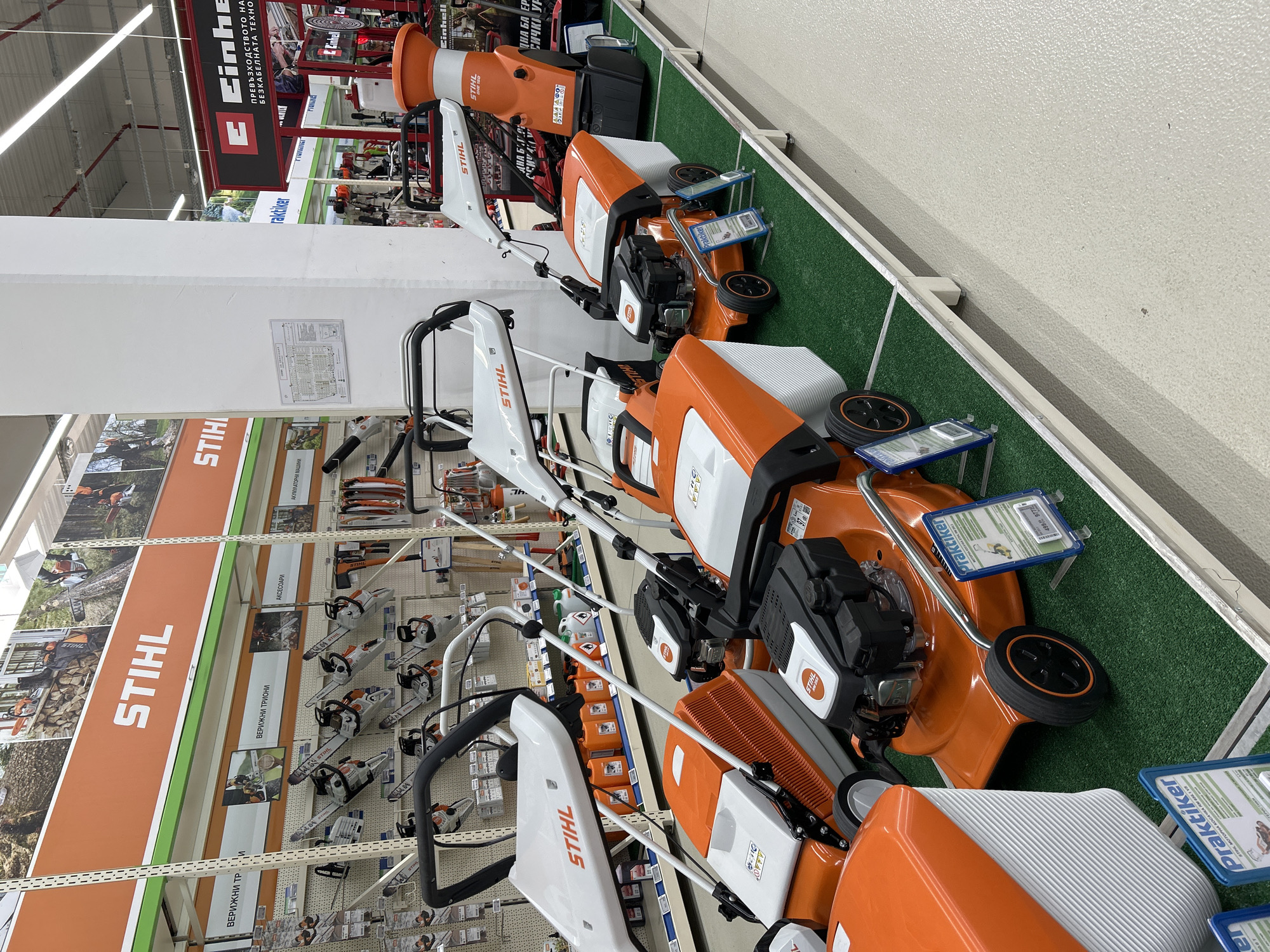 This is a vertically oriented photograph taken inside a retail store, likely a hardware or garden supply store, in Varna, Bulgaria. The primary subjects are Stihl branded lawnmowers and other gardening equipment displayed on shelves and on a green artificial turf floor. The scene showcases a well-organized retail environment with multiple products neatly arranged for sale.
Several orange and white Stihl lawnmowers are prominently featured in the foreground and middle ground, with their handles, wheels, and engines clearly visible. Some have blue and white price or information tags attached. In the background, the store's interior is visible, including shelving units stocked with various tools and equipment, as well as large Stihl promotional banners. One banner displays the Stihl logo and images of people using their products, along with text in Bulgarian, including "ВЕРИЖНИ ТР" (chainsaw), "ВЕРИЖНИ ТРИОНИ" (chainsaws), "АКСЕСОАРИ" (accessories), and "АКУЛГУЛАТОРНИ МАШИНИ" (battery-powered machines). Another banner mentions "ПРЕВЪЗХОДСТВОТО НА БЕЗКАБЕЛНАТА ТЕХНО" which translates to "The superiority of wireless technology." The overall impression is one of a modern retail space dedicated to outdoor power equipment. There are no people visible in the image. The lighting suggests it is daytime, likely during store operating hours.