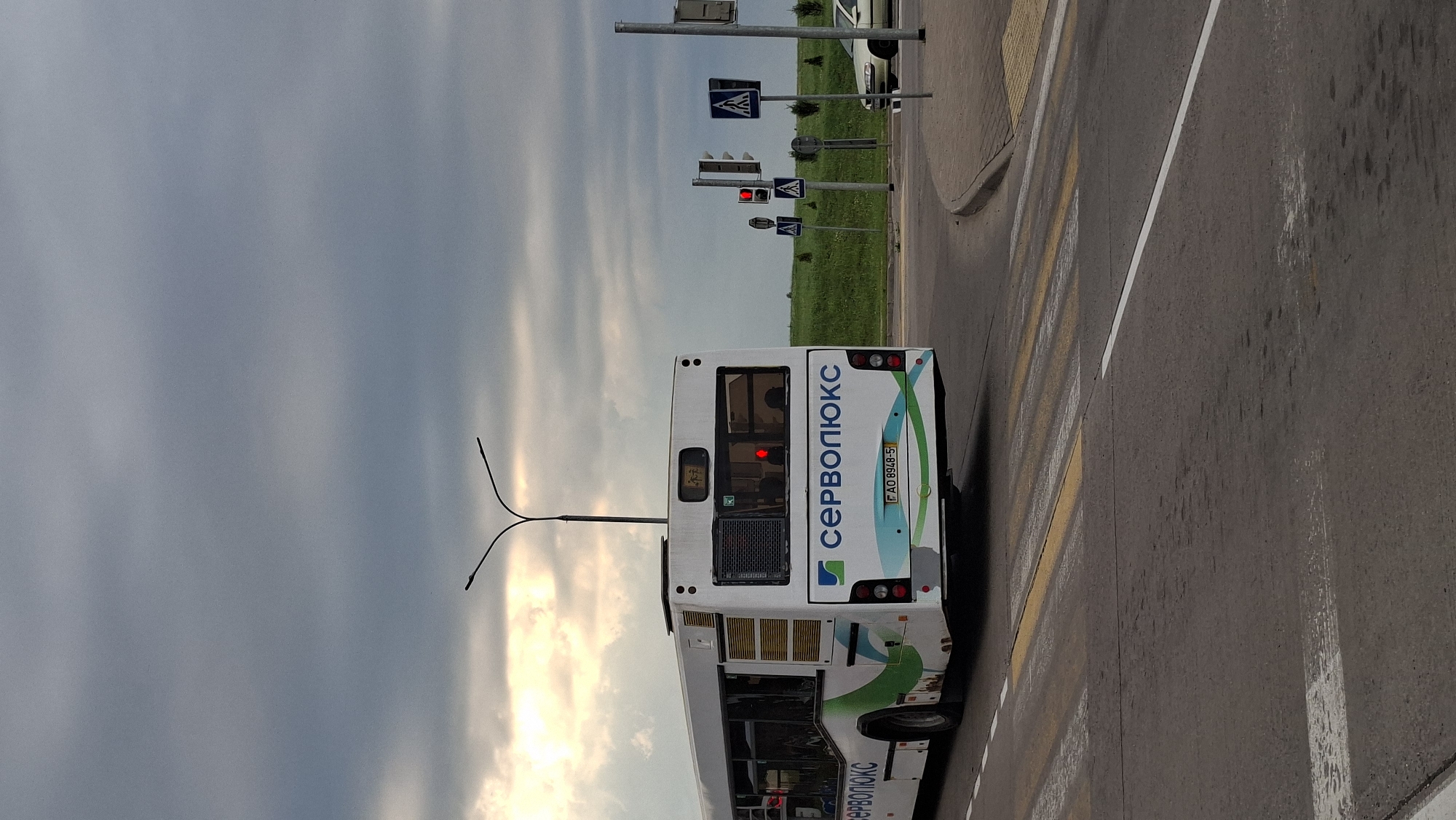 The image captures a rear view of a white bus with green and blue accents, bearing the Cyrillic text "СЕРВОПЛЮКС". The bus is on a paved road, approaching a pedestrian crossing marked with yellow and white stripes. In the background, a traffic light shows a red signal, and there are pedestrian crossing signs. A light-colored car is visible to the right, further down the road. The environment appears to be on the outskirts of a city, with a grassy embankment and a cloudy sky overhead. The image was taken in the city of Smaliavichy, Belarus.