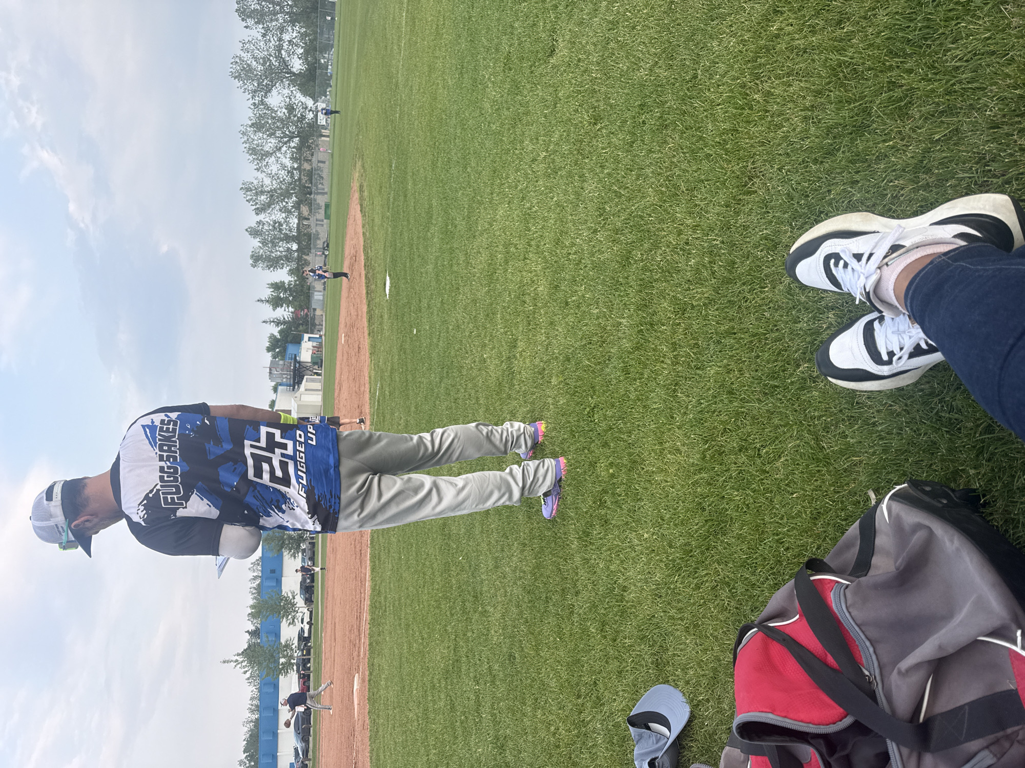 This image depicts a baseball field with a game in progress. In the foreground, a person's legs are crossed, wearing blue jeans and white sneakers with black accents. To their left is a duffel bag, partially red and gray with black straps, and a light blue baseball cap. In the mid-ground, a person wearing a gray baseball cap, a black and blue t-shirt with graphics, and gray pants stands looking at a clipboard. The t-shirt has "FUGG SAKES" and "FUGGED UP" printed on it. In the background, players are visible on the baseball diamond, with one player pitching. The field is surrounded by green grass and trees. Buildings and a chain-link fence are visible in the far background. The sky is overcast with scattered clouds. This scene is consistent with a sports field located in the city of Calgary, Canada.