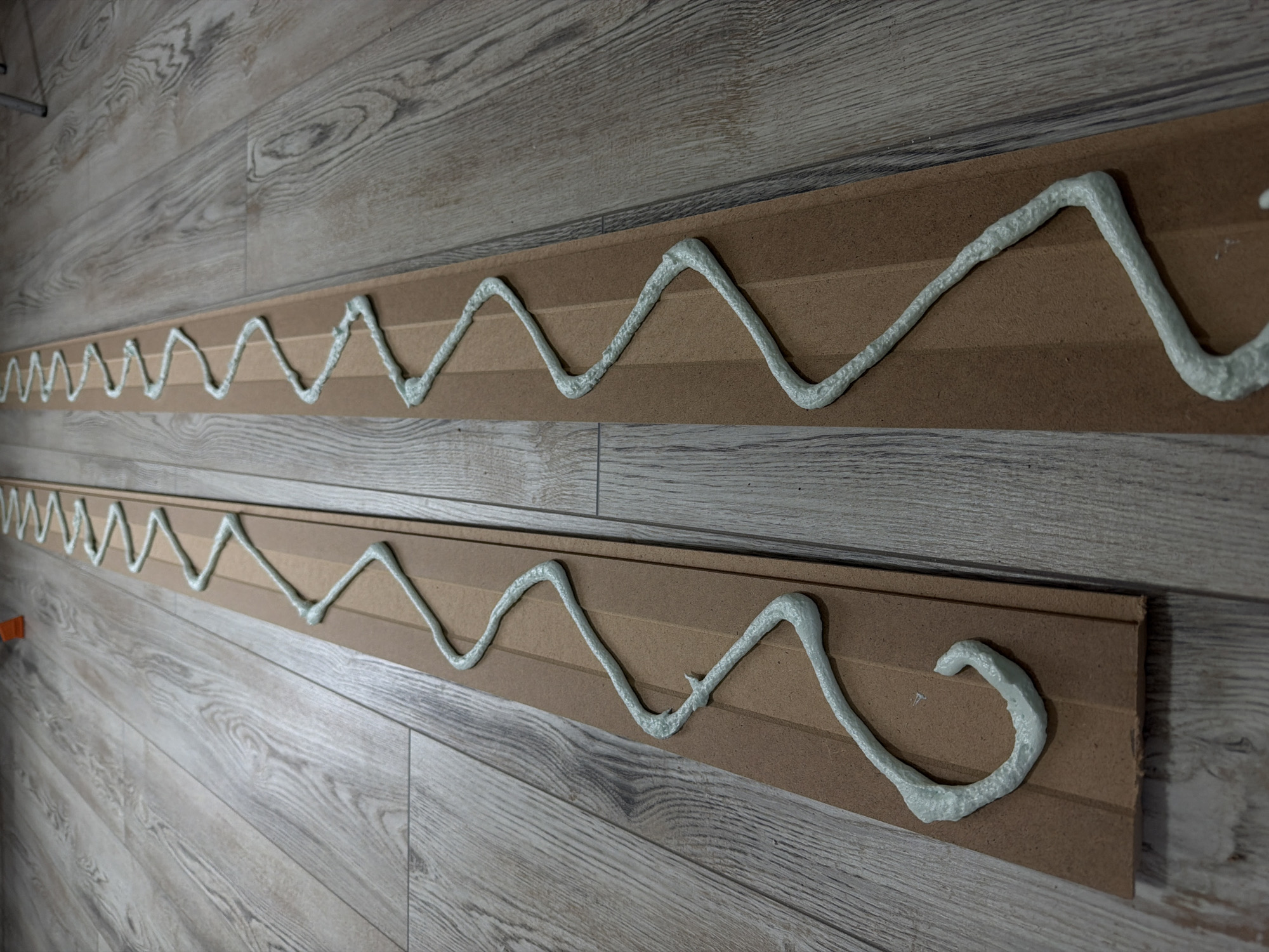 Two long, rectangular panels, likely made of light brown MDF or similar wood composite, are depicted lying flat on a light-colored wooden floor. Each panel features a thick, wavy bead of light blue-gray adhesive or sealant applied along its entire length. The adhesive appears freshly extruded, exhibiting a textured, somewhat frothy consistency.
The panels are positioned parallel to each other, running horizontally across the frame. The floor consists of planks with a distinct wood grain pattern in shades of light gray and brown, suggesting laminate or engineered wood flooring.
The scene captures a practical activity, likely the preparation of wall panels or molding for installation in an indoor setting. The adhesive is applied in a serpentine pattern, a common method to ensure good coverage and adhesion when mounting items.
No people are visible, and no specific time of day or weather conditions can be inferred, though the lighting is bright and even, typical of an indoor environment. There is no visible text. The location context for this activity is Dubovaya, Russia.