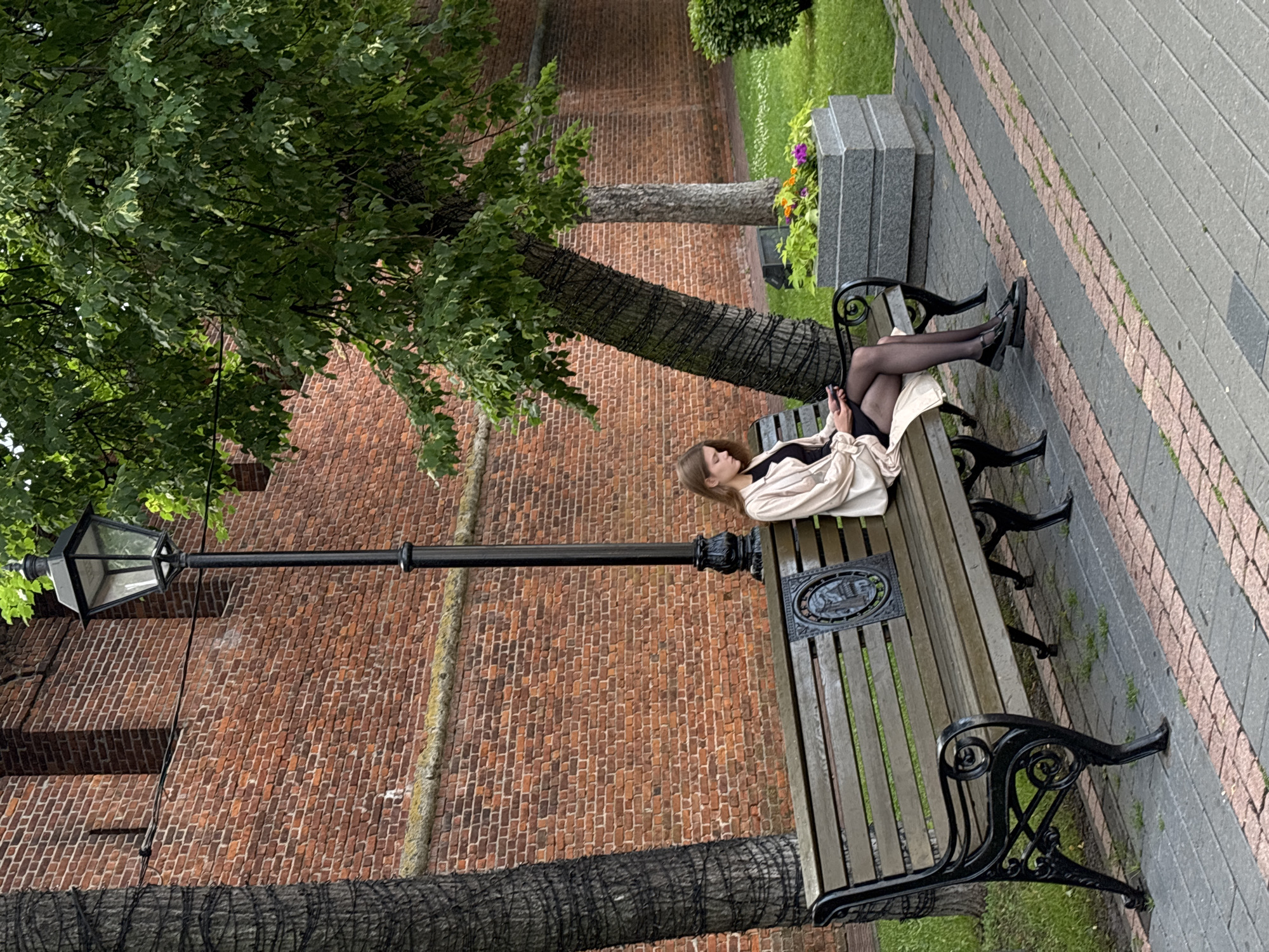 The image shows a young woman sitting on a park bench in Nizhny Novgorod, Russia. She is wearing black pantyhose, black shoes, and a light-colored jacket over black attire. She is looking at her phone, which she holds in her hands. The bench is made of wood and has black decorative metal legs. There is a decorative emblem on the bench. Behind the bench, there is a brick wall, and a lamppost is visible to the left. A large tree with green leaves is to the left of the bench, and its trunk is wrapped with wire or netting. The ground is paved with interlocking stones and some grass. In the background, there is a stone planter with flowers and more greenery. The scene appears to be a park or a public space with historical architecture. The lighting suggests it might be daytime, possibly overcast, as there are no strong shadows. The woman's posture and engagement with her phone suggest a moment of quiet leisure or waiting.
