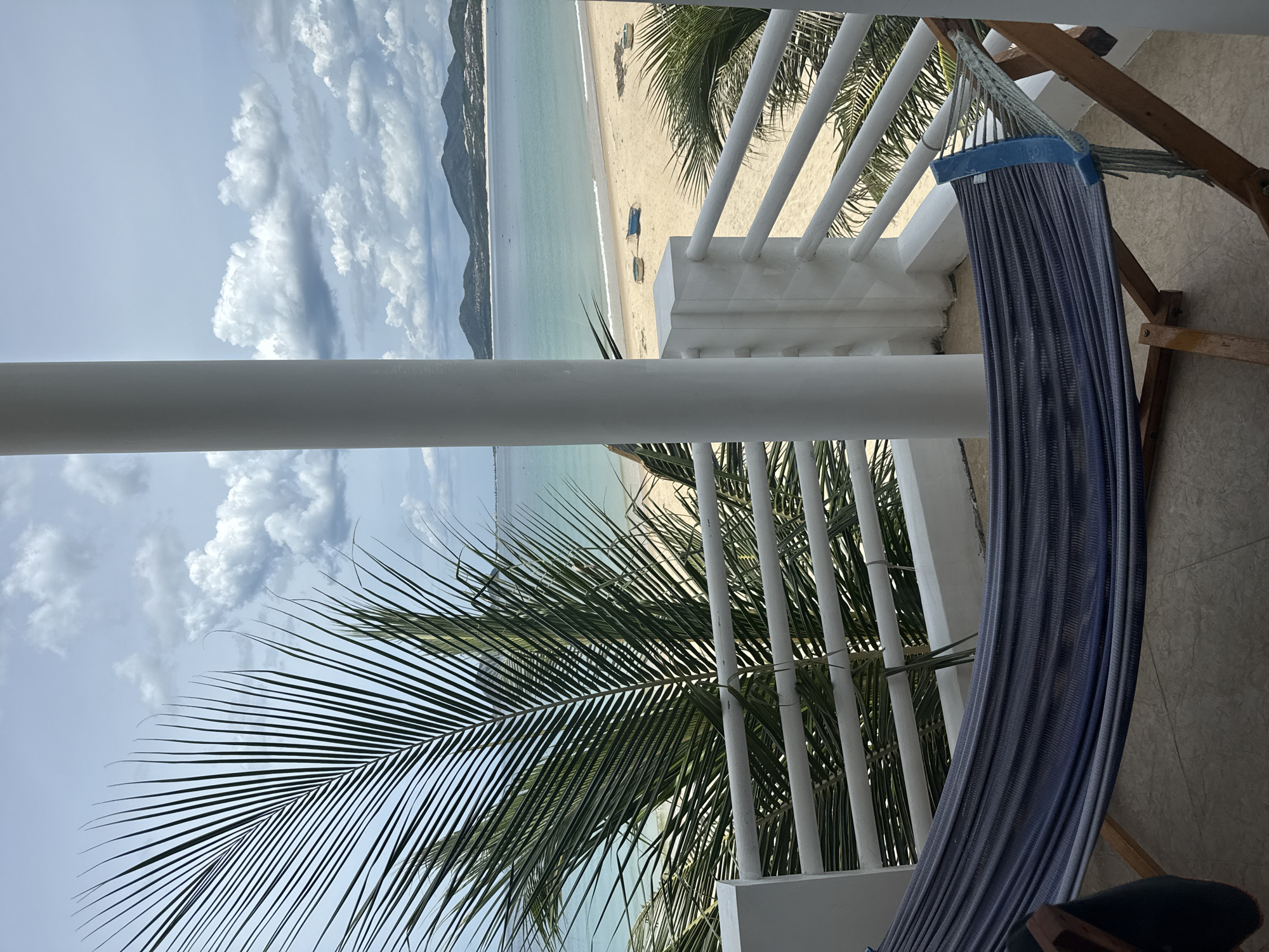 A hammock is positioned on a balcony overlooking a beach. The hammock is dark blue and grey with a wooden frame. A large palm frond hangs from the left side of the image, partially obscuring the view of the ocean and the beach. The beach is sandy, with a few small boats visible in the distance. The ocean is calm and turquoise, meeting a horizon line where faint, mountainous terrain is visible. The sky is cloudy with patches of blue. A white cylindrical pillar is situated in the foreground, slightly off-center to the right. The balcony has white railings.