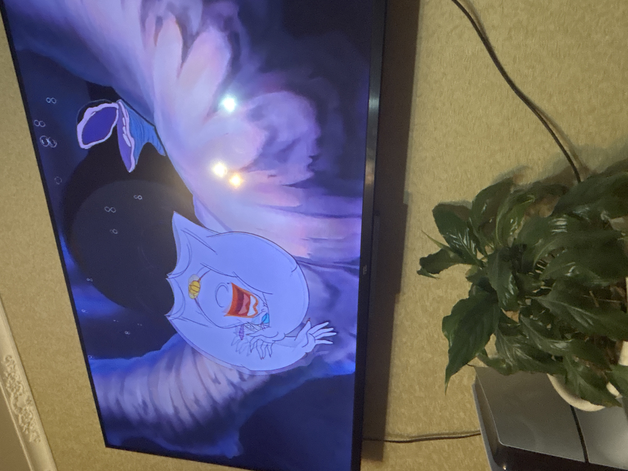 This is a photograph taken in Saratov, Russia, showing a television screen displaying an animated scene from Disney's "The Little Mermaid." The scene features Ursula, the sea witch, in her cavern, with bubbles floating in the dark blue water. She is depicted as a large, pale purple creature with tentacles, and her face is visible, showing a menacing expression with bright red lips and blue eyes. She is holding a large conch shell. In the foreground, a potted green plant sits on a surface below the television, with its leaves partially obscuring a white gaming console or electronic device. The wall behind the plant and television has a textured wallpaper. The lighting in the room is dim, with reflections visible on the television screen.