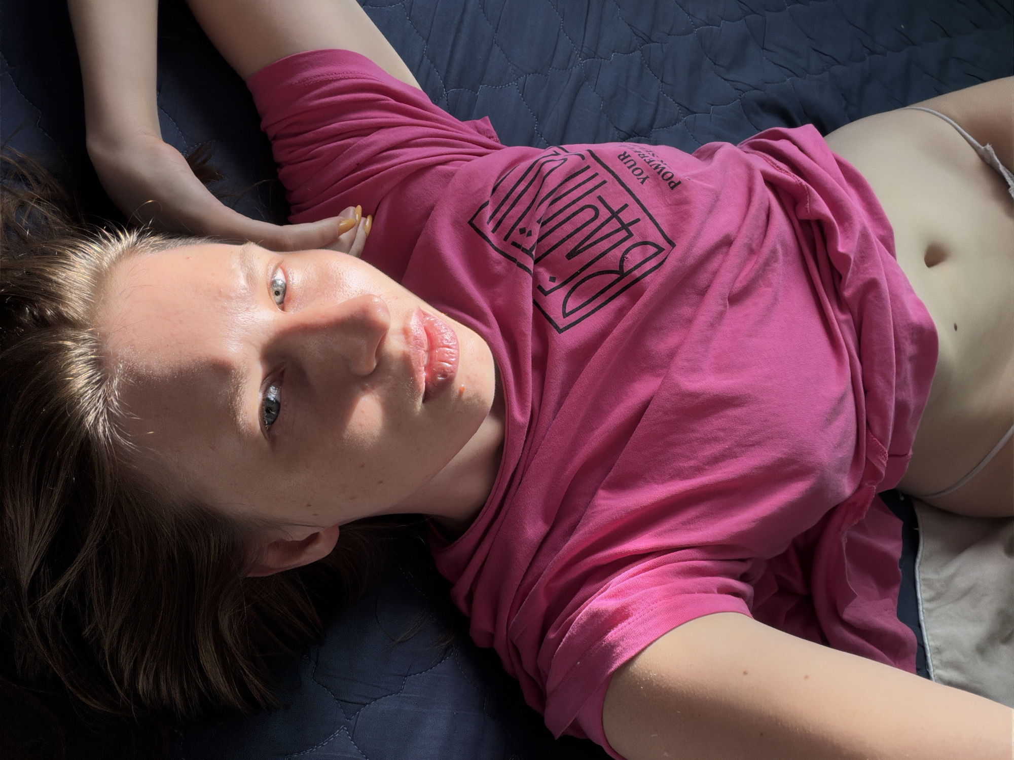 A young woman is lying on a dark blue quilted surface. She is wearing a bright pink t-shirt that is partially lifted, revealing her midriff and light-colored underwear. The t-shirt has a black graphic design on it with text. Her head is tilted back and to the left, with her right arm bent and resting behind her head. Sunlight casts shadows and highlights across her face and hair, which is brown and appears windswept. Her eyes are open and looking towards the camera. The overall environment suggests an indoor setting with natural light. The location is not specified within the city of Dongducheon-si, South Korea.