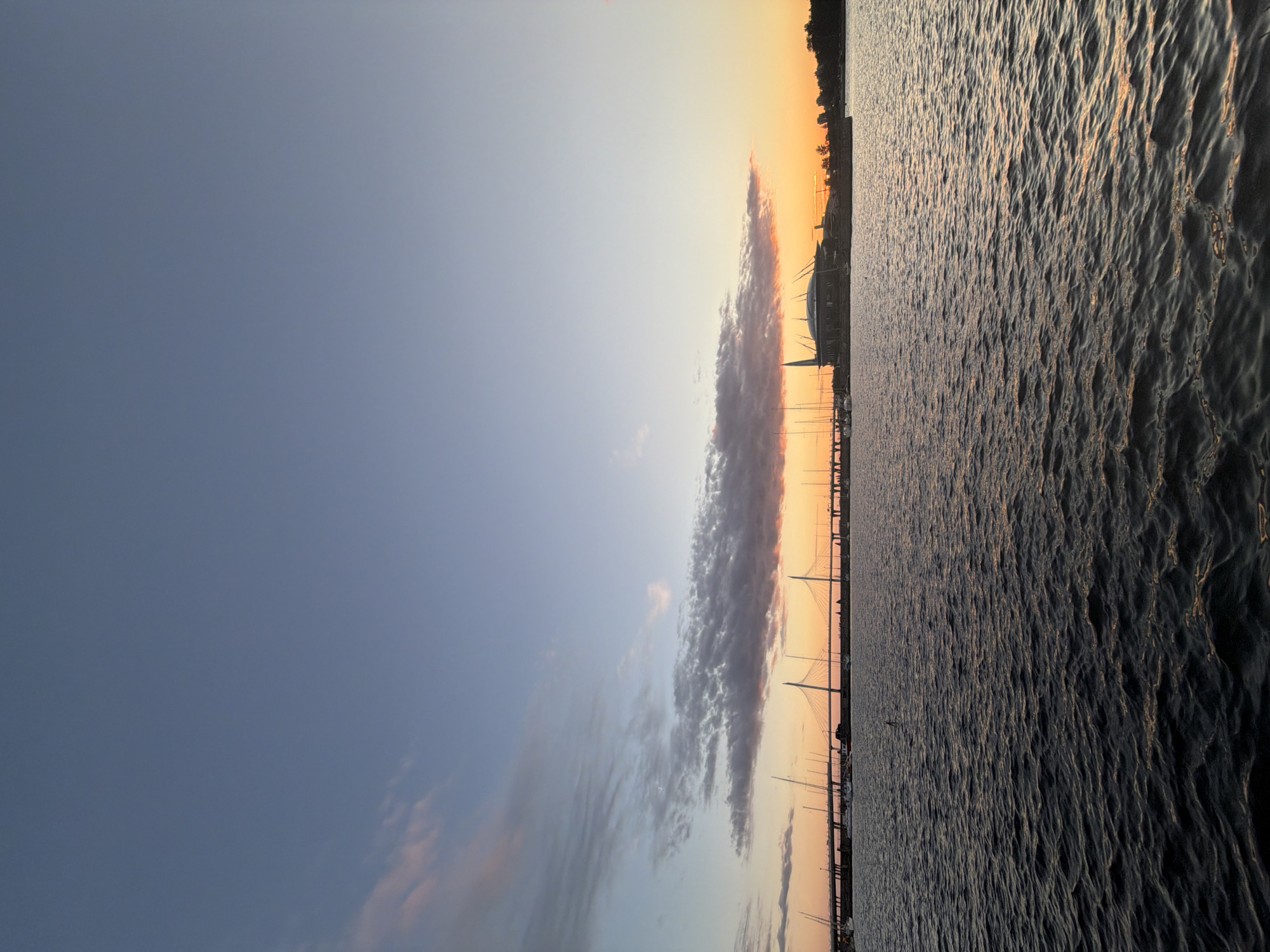 This is a horizontal, medium shot photograph taken from a watercraft, looking towards the shore at sunset. The water in the foreground is dark and choppy, reflecting the sky's colors. The sky transitions from a pale blue on the left to a vibrant orange and yellow on the right, indicating the sun is setting. A long, striated cloud stretches across the middle of the sky, catching the sunset's light.
On the shore, silhouetted against the bright sky, are various structures. A distinctive bridge with tall, angled towers spans across the water in the distance. Closer to the shore, there appear to be buildings, including one with a domed roof. Several masts of sailboats are visible along the waterline, suggesting a harbor or marina. Trees line the far edge of the land.
The scene evokes a sense of calm and beauty, typical of a serene sunset over a body of water. The time of day is clearly sunset. The location is implied to be Saint Petersburg, Russia, with the prominent bridge being a recognizable landmark. There is no visible text in the image.