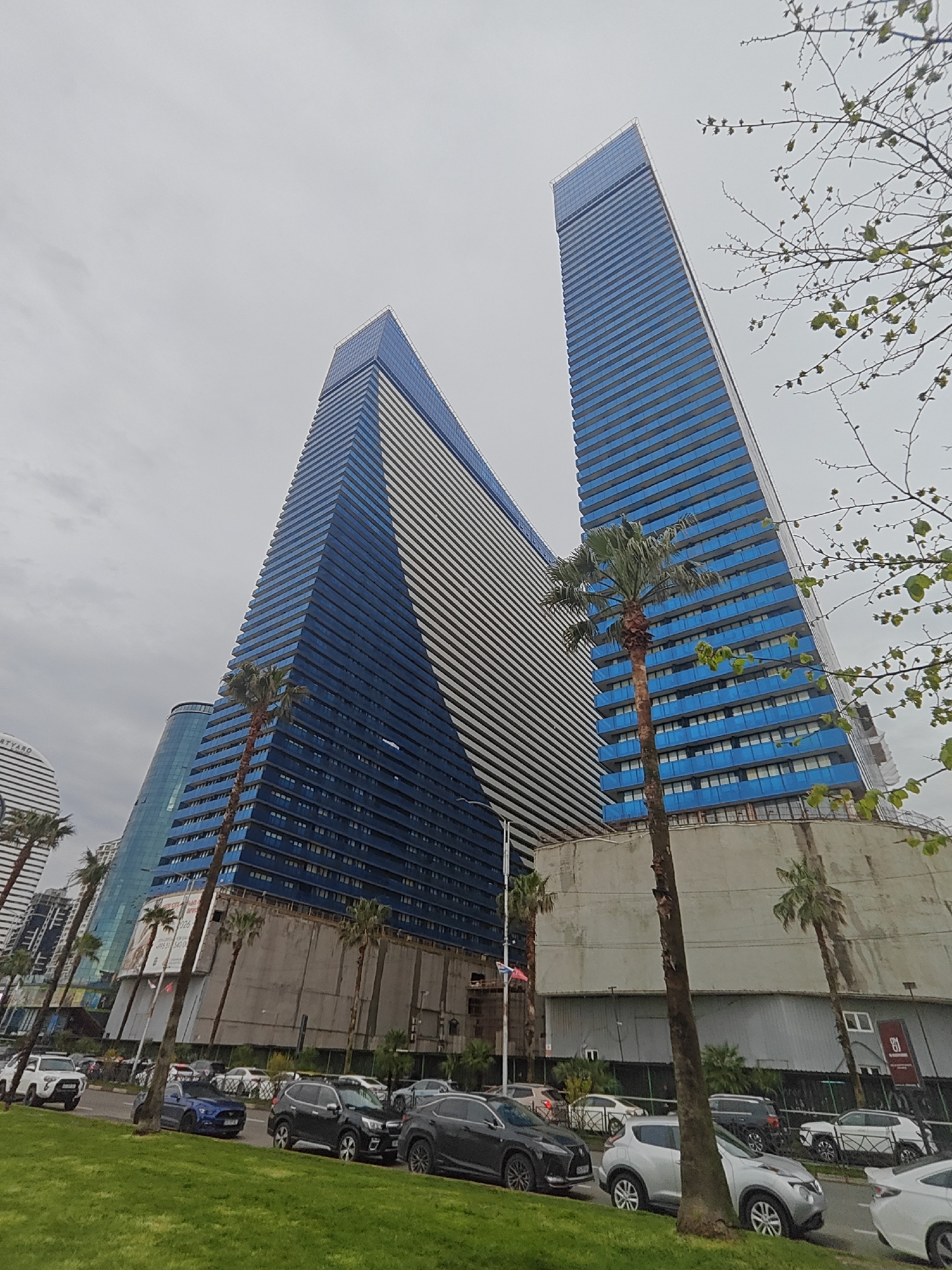 The image captures a vibrant urban scene dominated by modern architecture under an overcast sky. Two towering, sleek skyscrapers with distinctive blue and white/grey striped facades are the central subjects, with the taller one positioned on the right. Numerous palm trees are interspersed throughout the foreground and midground, lining the street and standing in front of the buildings, indicating a warm climate.
The scene depicts everyday city life, with a paved road occupied by a variety of parked and moving vehicles, including several SUVs, sedans, and a white pickup truck. A green grassy median separates the road from the foreground.
Notable details include the overcast, daytime sky, suggesting a cloudy day. In the background on the left, a curved white building bears the partially visible text "COURTYARD," likely indicating a Courtyard by Marriott hotel. A flagpole with a small red and white flag is also visible. At the base of the right skyscraper, signs of ongoing construction or renovation are present, including a large banner with partially visible text that includes "TEL.: 026" and other obscured words. Further to the right, a red sign with a stylized white "8" or similar logo is partially visible near some construction barriers. The overall impression is one of a modern, developing city environment.