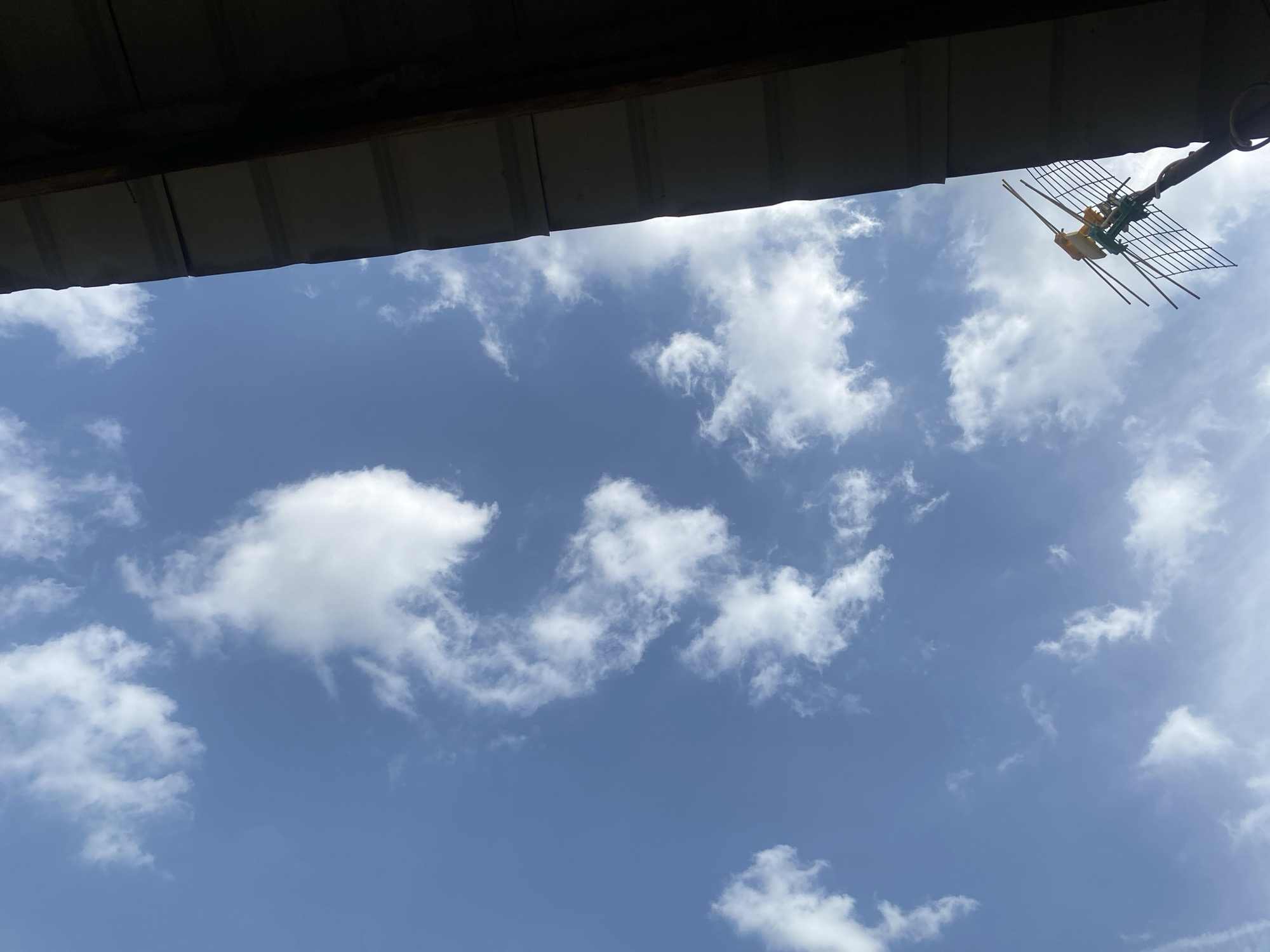 The image presents a bright, daytime view of a clear blue sky adorned with scattered white clouds, captured from beneath a dark overhang. The overhang, likely the underside of a roof or eave, made of corrugated material, occupies the top portion of the frame and is cast in shadow.
In the upper right corner, an outdoor television antenna is visible, featuring a metallic grid, several director elements, and distinctive yellow and green components, mounted on a pole. The clouds are fluffy cumulus formations, suggesting fair weather. No people or discernible activities are depicted, and there is no visible text. The scene is a common outdoor setting, possibly in a residential area, consistent with the provided location context of Lagos, Nigeria.