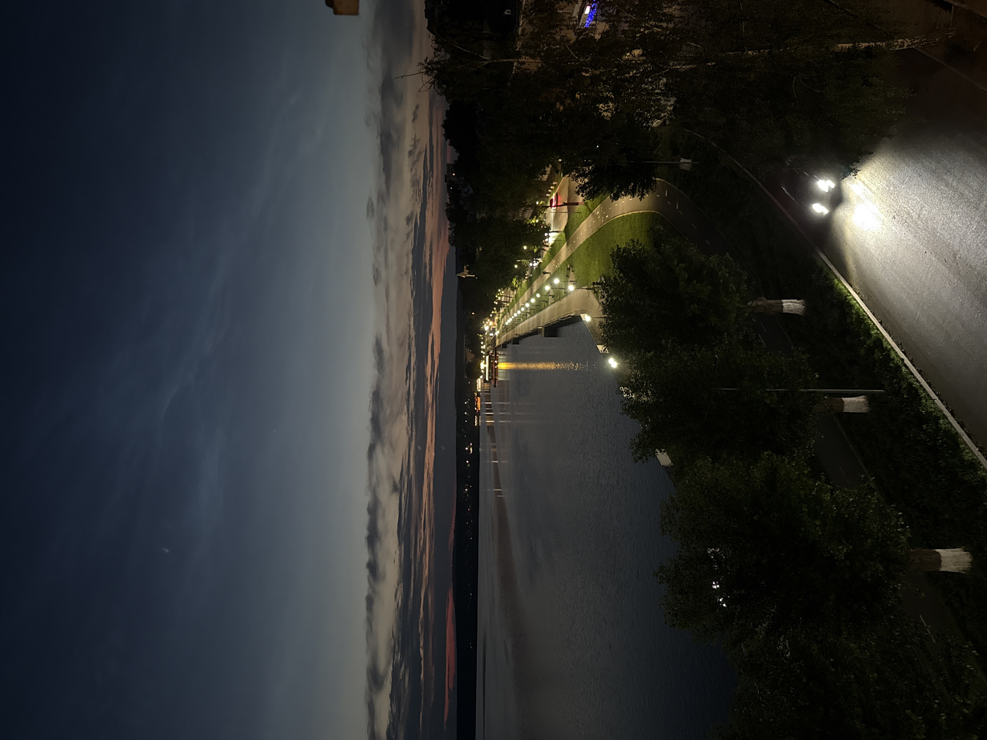 This is a nighttime aerial view of a waterfront promenade in Votkinsk, Russia. The scene is dominated by a large body of water on the left, reflecting the lights from the promenade and distant buildings. The promenade itself is lined with trees and illuminated by numerous streetlights, creating a warm glow. A paved pathway runs alongside the promenade, with a road to its right. A single car with its headlights on is visible on the road, its beams illuminating the asphalt. The sky above is dark, with subtle hints of clouds and the fading colors of sunset. The distant shoreline features a line of buildings and trees. The overall atmosphere is serene and tranquil.