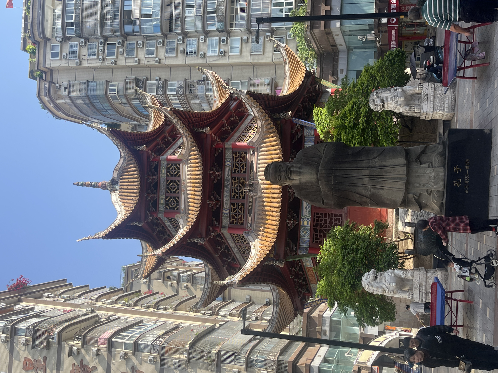 This vibrant daytime image captures a bustling urban scene in Jiangjin, China, combining traditional Chinese architecture with modern development. The central focus is an ornate, multi-tiered pagoda-style building with curved, golden-tiled roofs and intricate red and dark green decorative elements. In front of this structure stands a large, dark stone statue of a man, identified by the inscription on its base as "孔子" (Kǒngzǐ - Confucius), with the dates "生于公元前551—前479" (born in 551 BC - 479 BC).
Flanking the statue are two large, white stone lion statues, typical guardians of Chinese temples and important buildings. Lush green trees and bushes are visible around these statues and near the traditional building, providing a touch of nature.
The background is dominated by tall, modern apartment buildings with numerous windows, balconies, and air conditioning units, showcasing the dense urban environment. Some balconies feature small plants or clothes hanging. The sky is clear and bright blue, indicating sunny weather.
Several people are seen in the foreground and midground. One man in a green and white striped shirt stands near a red and blue table. Another person is pushing a baby stroller, while two individuals walk in the immediate foreground, one wearing a black jacket. Other blurry figures suggest a lively public space.
Visible text on a red sign in the background includes characters that appear to be part of a business name, possibly "金盛..." (Jīn Shèng...). The scene portrays a common street or public square, where daily life unfolds against a backdrop of cultural heritage and contemporary urbanism.