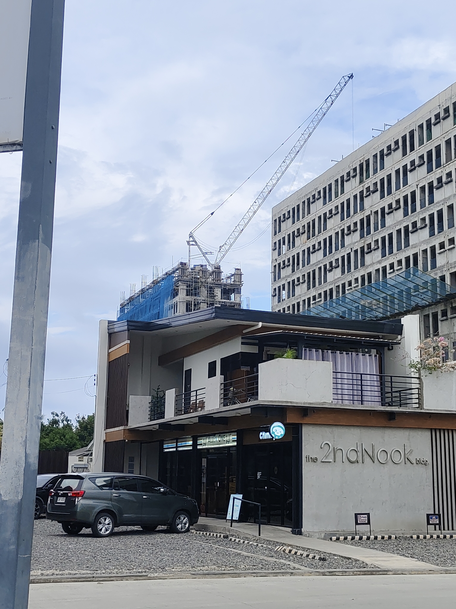 A construction crane is visible in the background, extending over a building that appears to be under construction, wrapped in blue netting. In the foreground, a modern, multi-story building with a glass walkway and balconies is shown. The building has a sign that reads "the 2ndNook bldg.". Several vehicles are parked in the gravel lot in front of the building, including a dark grey SUV. The sky is cloudy.