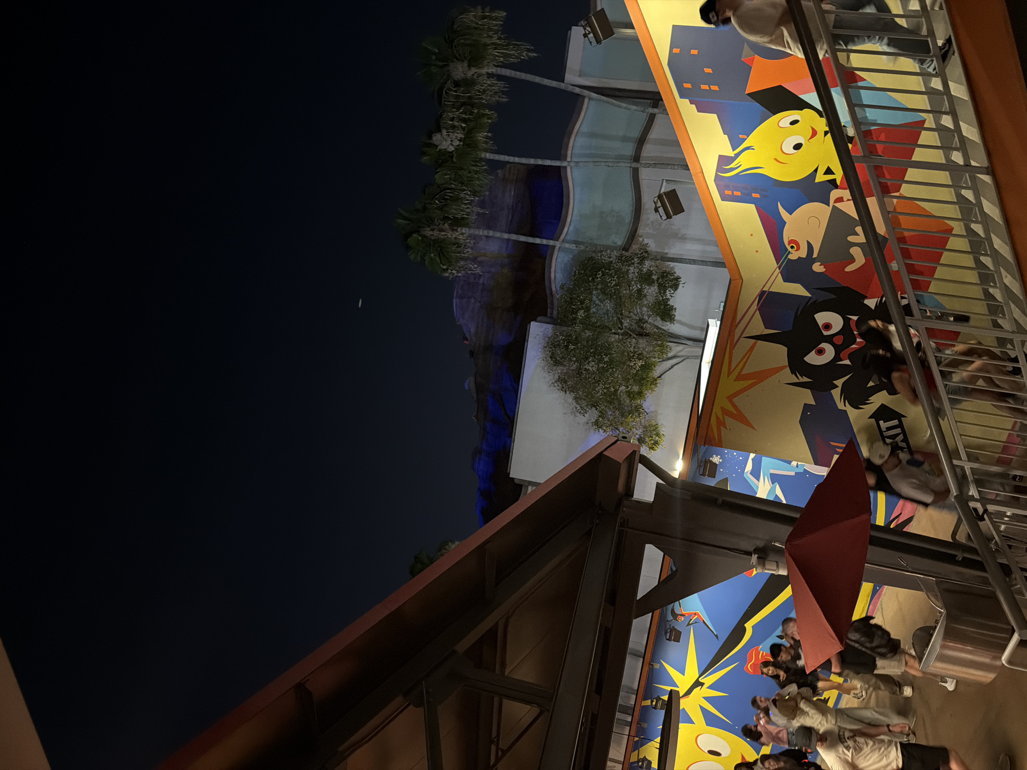 The image shows a nighttime scene at a location in Anaheim, USA. Tall palm trees are visible in the mid-ground, illuminated with blue light. Cartoon character themed building with a yellow character with hair that is yellow. The foreground reveals a partially visible metal and wood structure. An open red umbrella is also in the foreground. People are walking around in the background, including some that are standing under the umbrella. A railing and stairs are running along the side. The sky is dark, with a small crescent moon visible in the upper left.