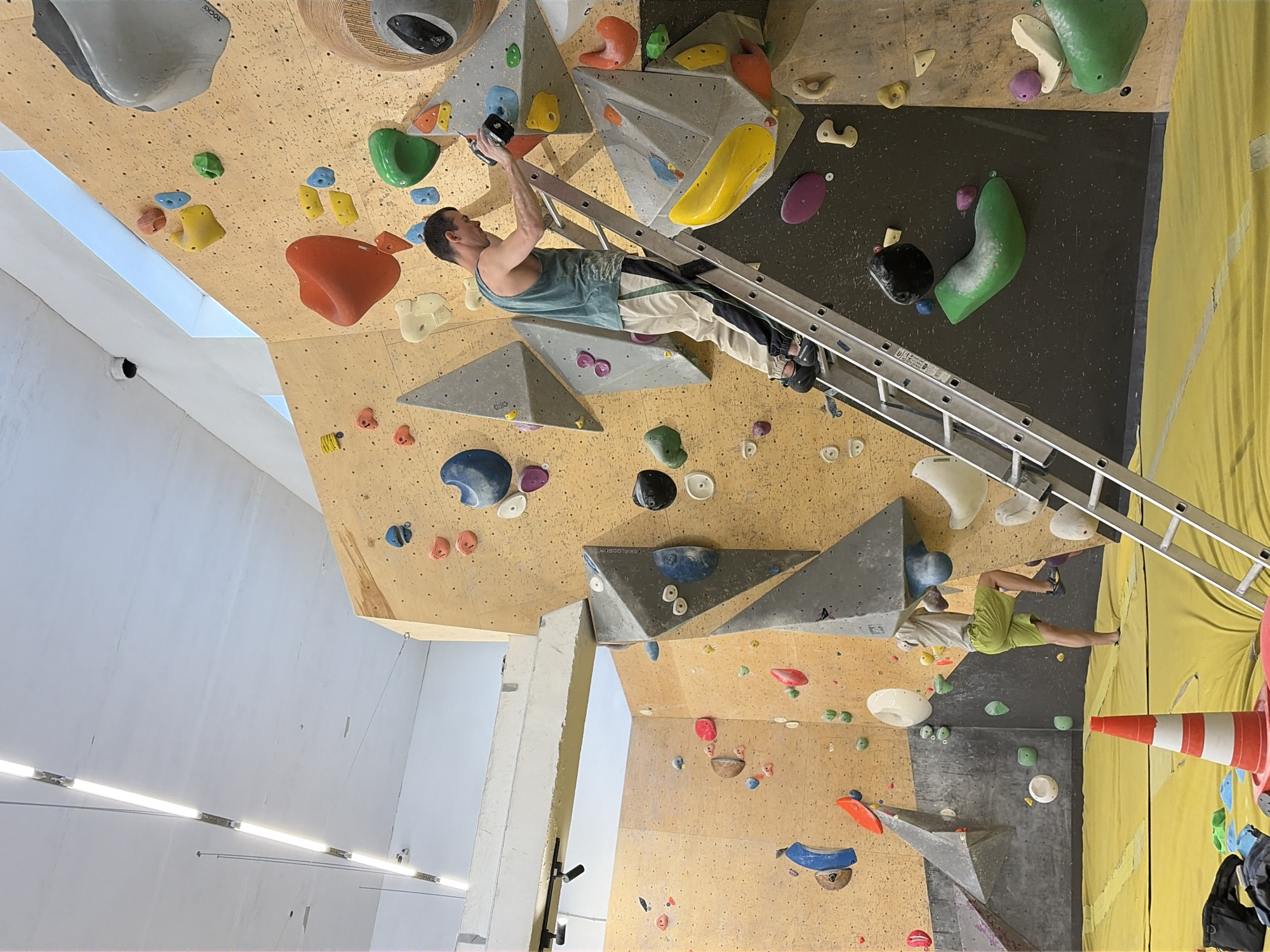 The image depicts an indoor bouldering gym. A man in a teal tank top and beige pants with black and green stripes is ascending a silver ladder placed against a large, yellow climbing wall. He is holding a black object in his right hand and appears to be reaching for a climbing hold with his left hand. His shoes are black. To the left of the ladder and further down the climbing wall, another person is visible, wearing green shorts and a light-colored shirt. They are standing on a yellow mat, with their right leg bent as if in the middle of a climbing move. The climbing wall itself is covered with various colorful holds and geometric structures. The gym is well-lit with fluorescent lights on the ceiling. A bright orange and white traffic cone is placed on the yellow mat in the foreground. The overall environment is that of an active recreational space.