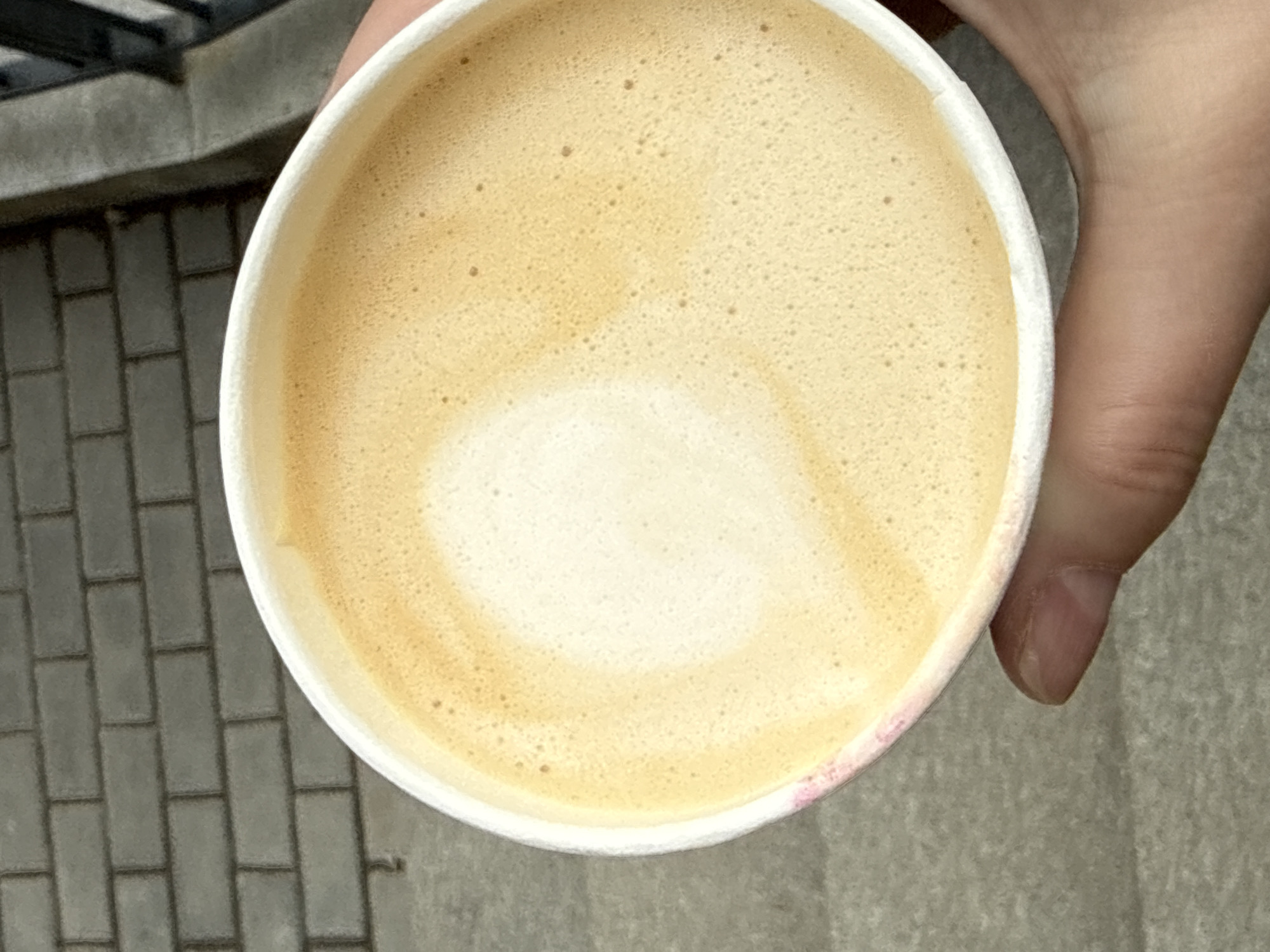 A close-up, top-down shot depicts a white paper cup filled with a latte. The latte has a creamy, light brown surface with a lighter, central area, suggesting latte art. The cup is held in a hand, with a thumb visible on the right side of the image. The background features a paved area with light grey bricks in the lower-left and a light grey surface on the right. The scene appears to be outdoors. There is no indication that the scene is in Izhevsk, Russia.