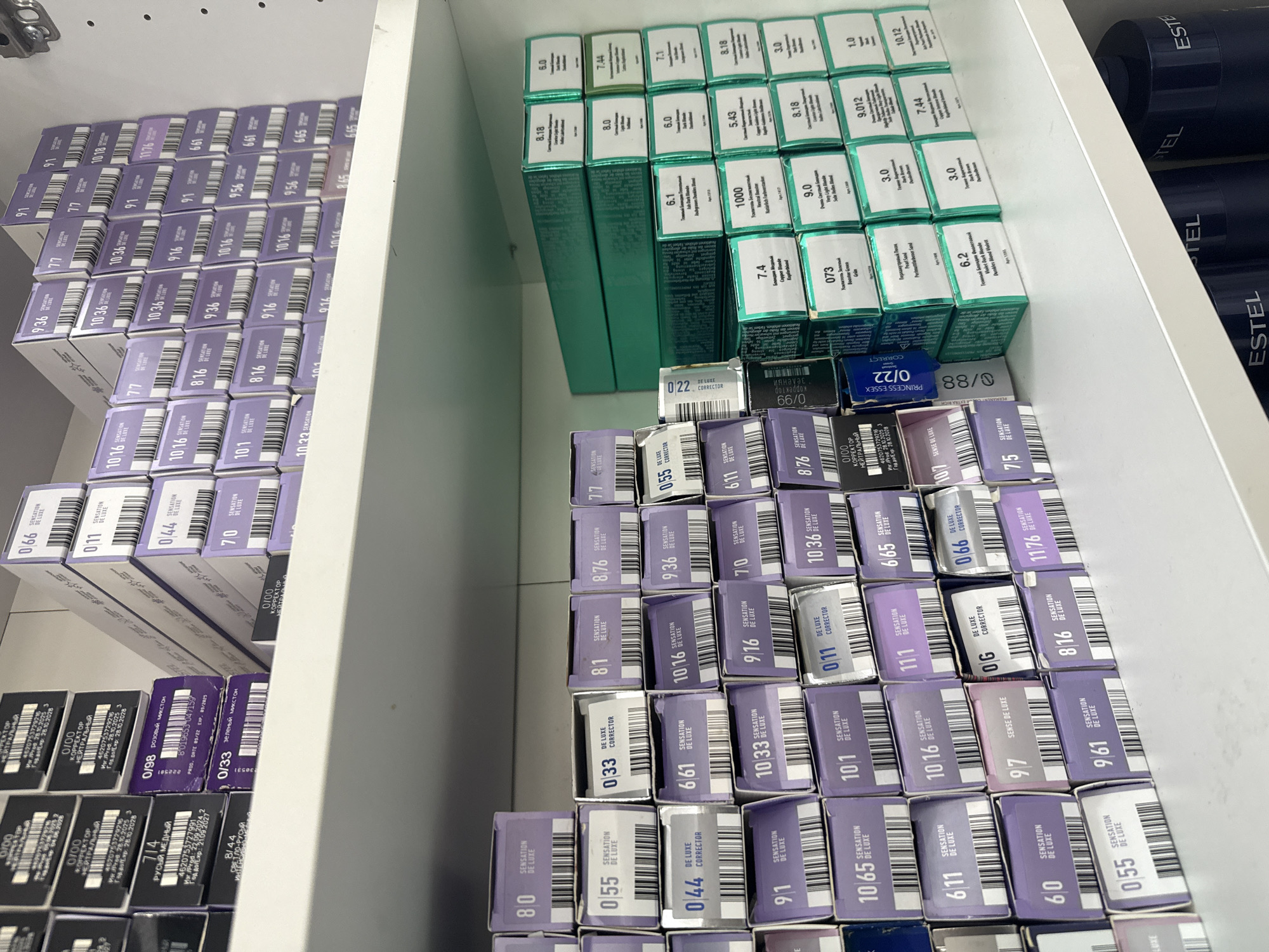 This is a photograph of a drawer containing hair dye boxes, arranged neatly in rows. The boxes are predominantly purple, green, and black. The brands visible are "Estel" and "Sensation De Luxe." The location is implied to be Chaikovsky, Russia, due to the presence of Cyrillic text on some of the boxes.