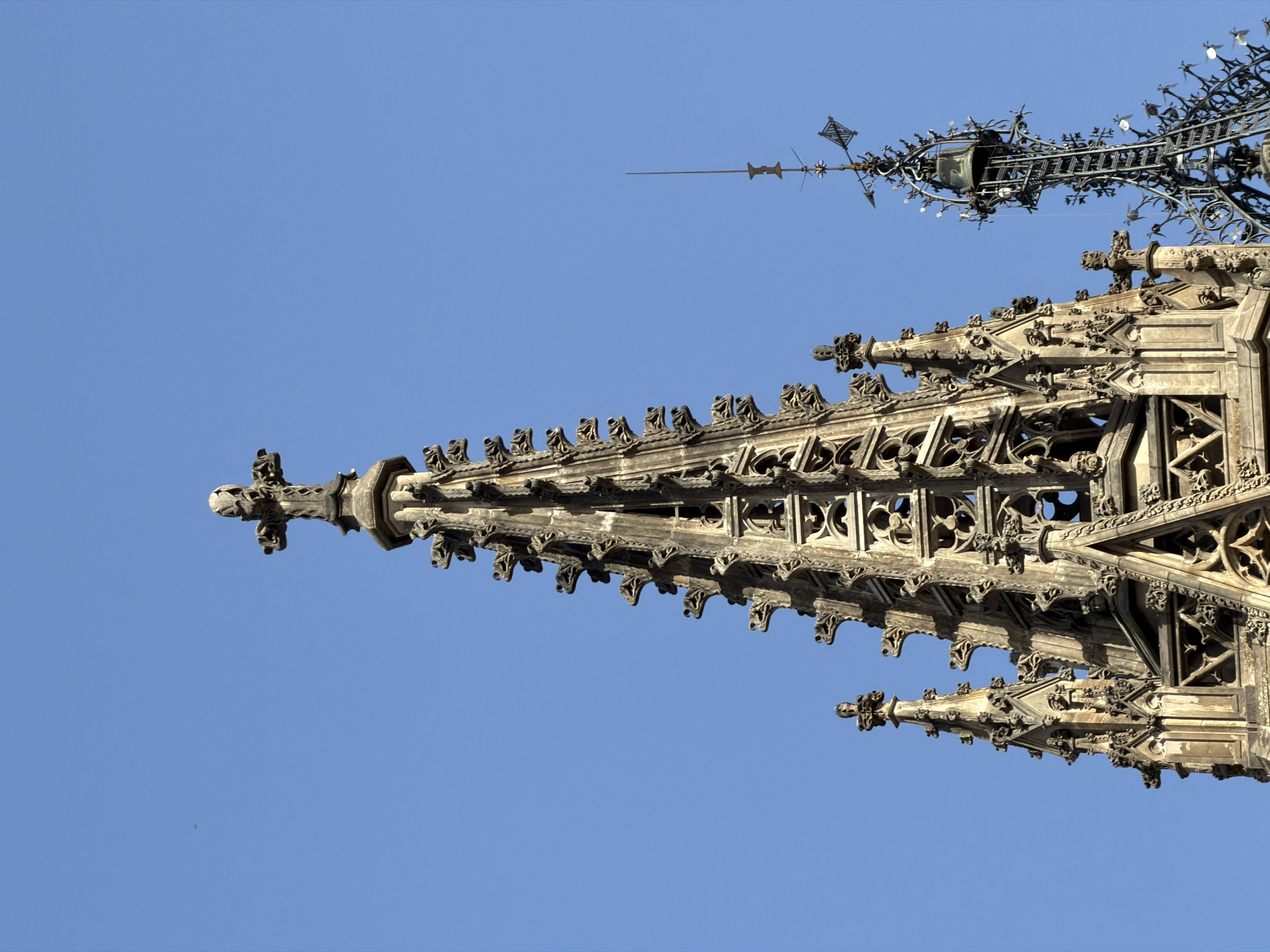 The image shows the spires of a Gothic cathedral against a clear blue sky. The main spire in the foreground is made of stone, with intricate carvings and decorative elements such as gargoyles. It culminates in a cross. To the right, a second, taller spire is visible, made of darker metalwork with a bell and a weather vane at its apex. The scene is likely from Barcelona, Spain, given the architectural style commonly found in its historic cathedrals.