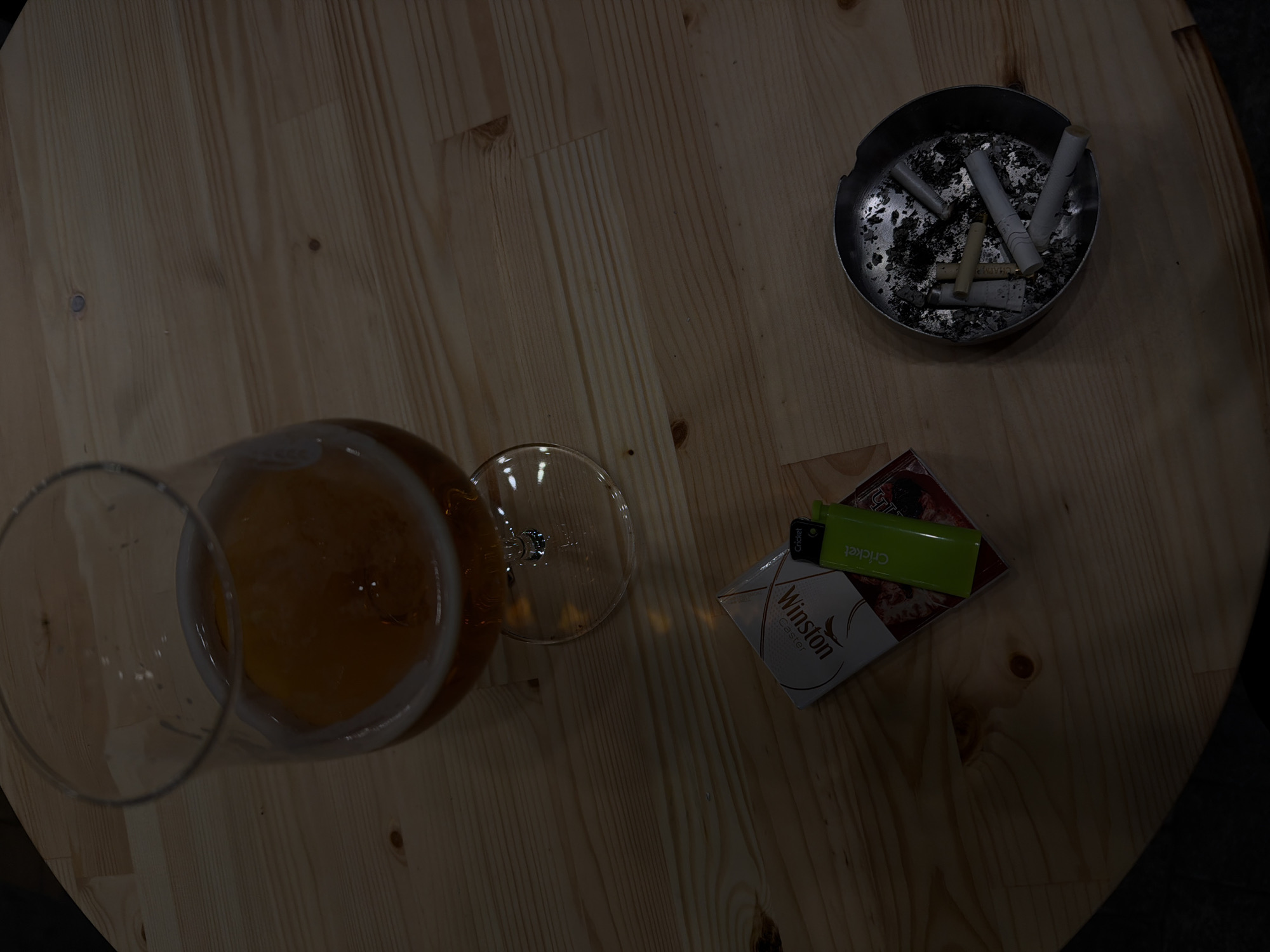 The image depicts a wooden table, likely in a pub or bar setting. There is a glass of beer, an ashtray filled with cigarette butts, a pack of Winston cigarettes, and a green Cricket lighter. The location, Yerevan, Armenia, is not evident from the image itself, but might be assumed if such information is given. No people are visible. The image appears to be a candid shot of a table setting, suggestive of a casual, perhaps social, environment.