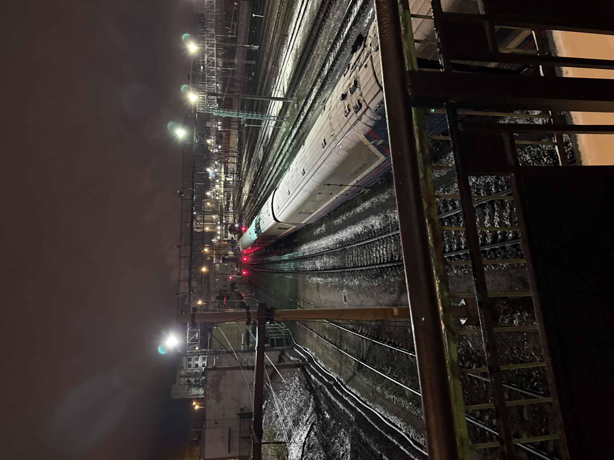 The image captures a nighttime scene at a train station in Moscow, Russia. A train is situated on the tracks, partly covered in what appears to be snow or frost. Overhead lighting illuminates the railway infrastructure. The perspective is from an elevated position, looking down onto the train and tracks, with a metal railing or structure partially obscuring the view. The sky is dark and overcast. Buildings are seen in the distance.