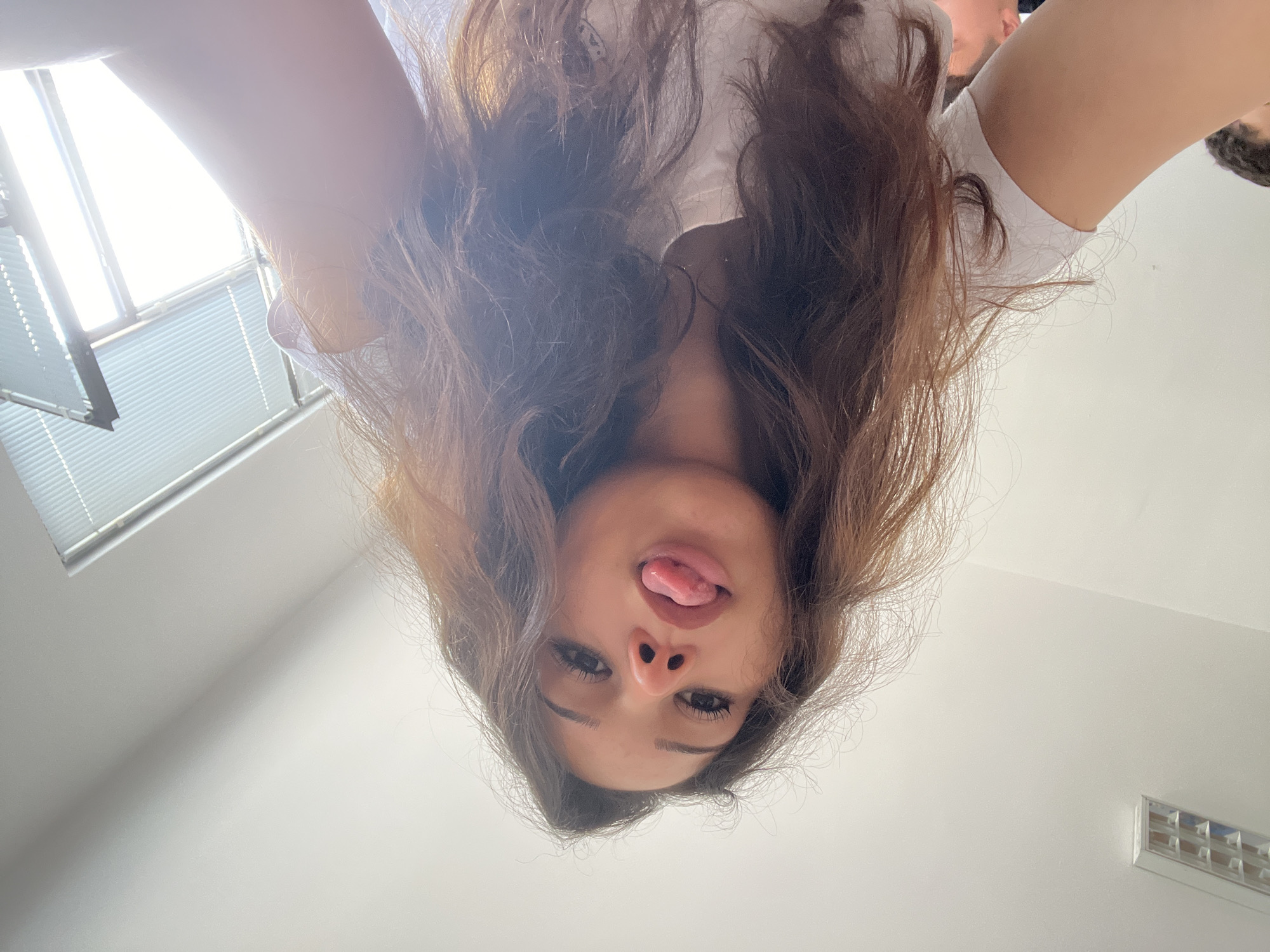 The image captures a low-angle shot looking upwards at a person, likely female, with long, wavy brown hair, positioned directly above the camera. Her face is presented upside down from this perspective, with her tongue playfully sticking out, suggesting a candid or humorous moment, possibly a selfie. She has dark eyes and is wearing a white top.
The background reveals an indoor setting. To the left, a brightly lit window with horizontal blinds is visible, indicating daylight. This strong light causes some glare in the upper left quadrant. The walls and ceiling are plain white. In the upper right corner, a small portion of another person's head or hair can be seen. Further right on the ceiling, a rectangular light fixture with a grid pattern is visible. The scene suggests a modern, well-lit interior, possibly a home or office. No text is discernible in the image. The overall mood is playful and lighthearted due to the subject's expression.