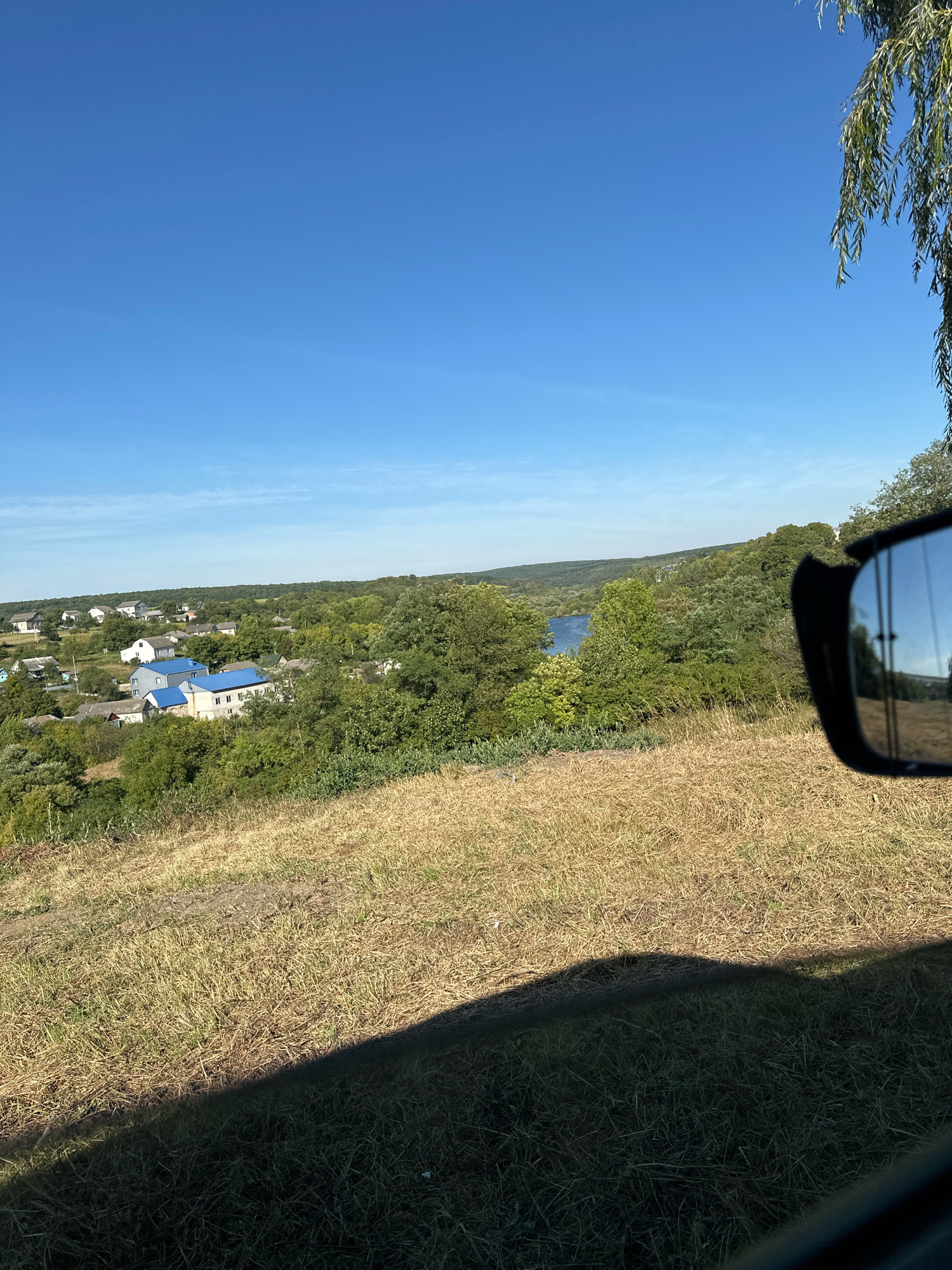 An exterior, eye-level, full shot captures a rural landscape with a clear blue sky and a distant view of a village. In the foreground, dry, golden grass covers a sloping hill, casting a dark shadow across the bottom of the frame. To the right, a car's side mirror reflects the scene. Beyond the grassy hill, a cluster of houses with blue roofs is visible, nestled among lush green trees. Further in the distance, a body of water, likely a river or lake, is seen, surrounded by more trees and rolling hills. The scene is set in the city of Makiv, Ukraine.