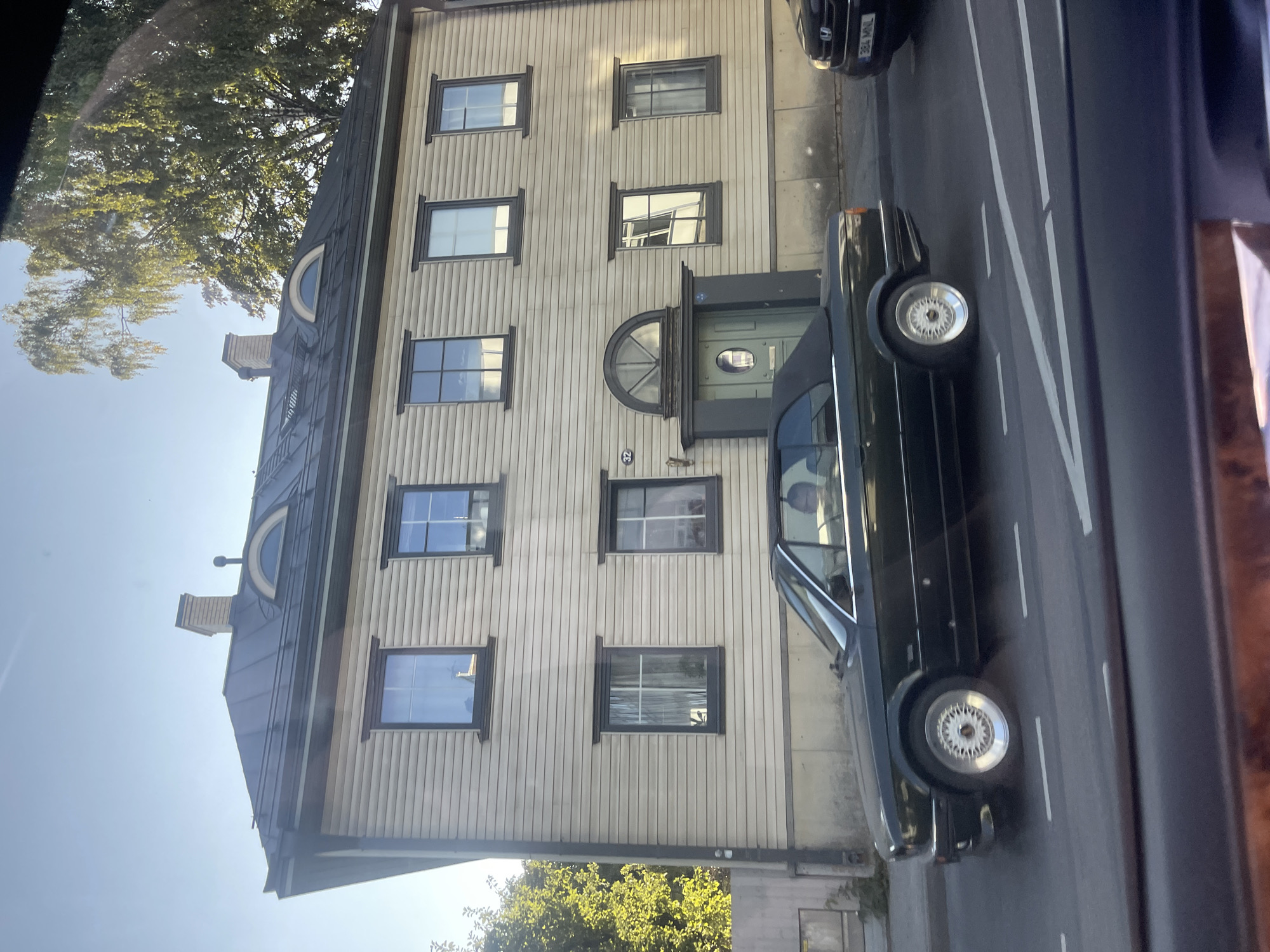 A dark green convertible BMW E30 is driving on a paved road in front of a light-colored wooden building. The building has a dark roof with dormer windows and a prominent chimney. The convertible top of the BMW is down, and a person is visible in the driver's seat. The car has chrome trim and distinctive white multi-spoke alloy wheels. The road has white lane markings. The scene is illuminated by natural sunlight, suggesting it is daytime. Trees with green foliage are visible to the left and right of the building. A black Honda CR-V is parked on the right side of the road behind the BMW. The license plate of the Honda is visible and reads "384 MNL". The overall environment suggests an urban or suburban setting, likely in Tallinn, Estonia, given the architectural style of the building and the presence of the car.