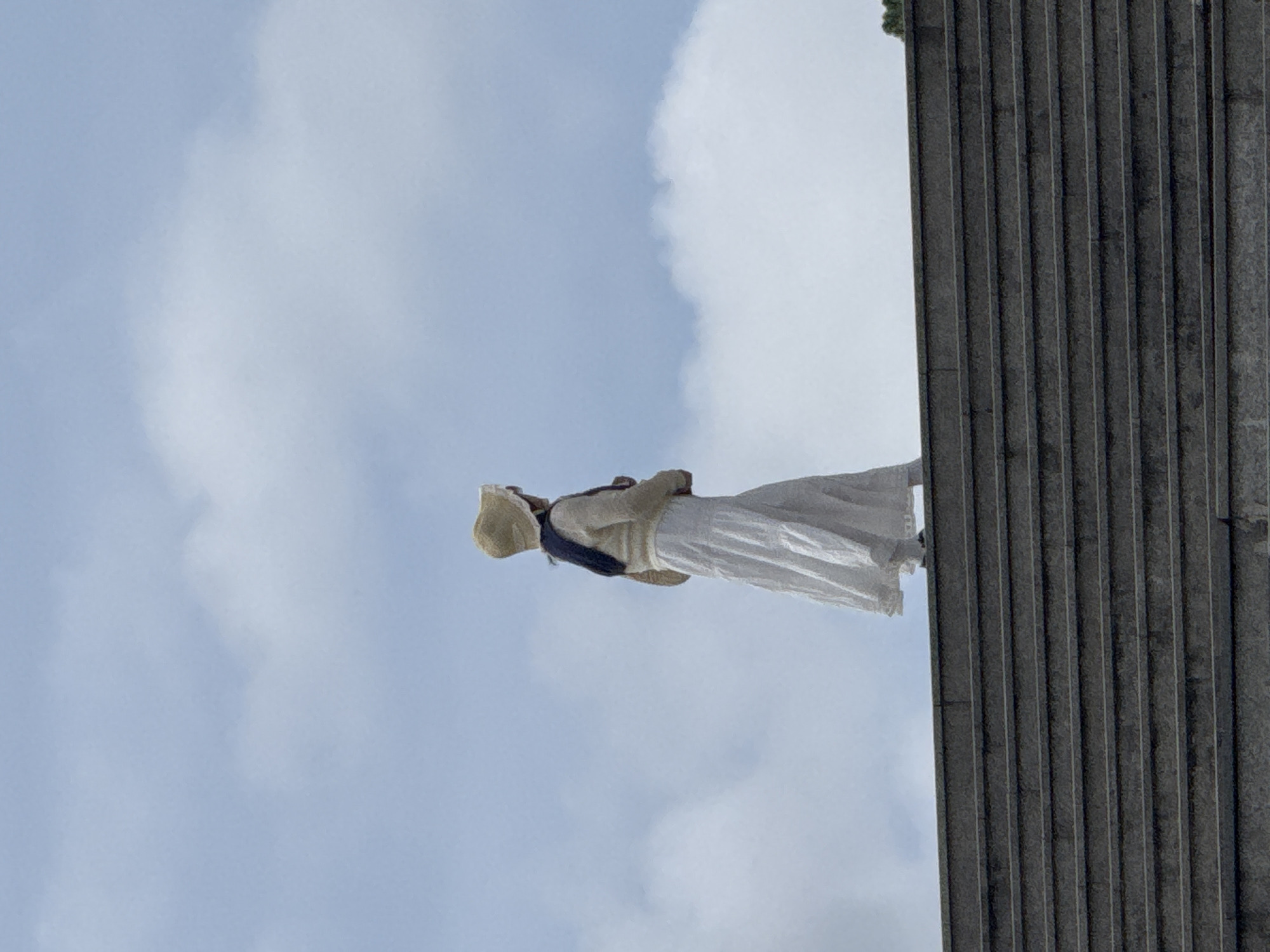 A person wearing a straw hat and a long white skirt stands on a set of stone stairs, looking out at the sky. The person is wearing a light-colored sweater over a dark shirt or scarf. The sky is cloudy, with patches of blue visible. The stairs are wide and made of dark gray stone. The scene appears to be outdoors. The image is taken from a low angle, looking up at the person and the sky.