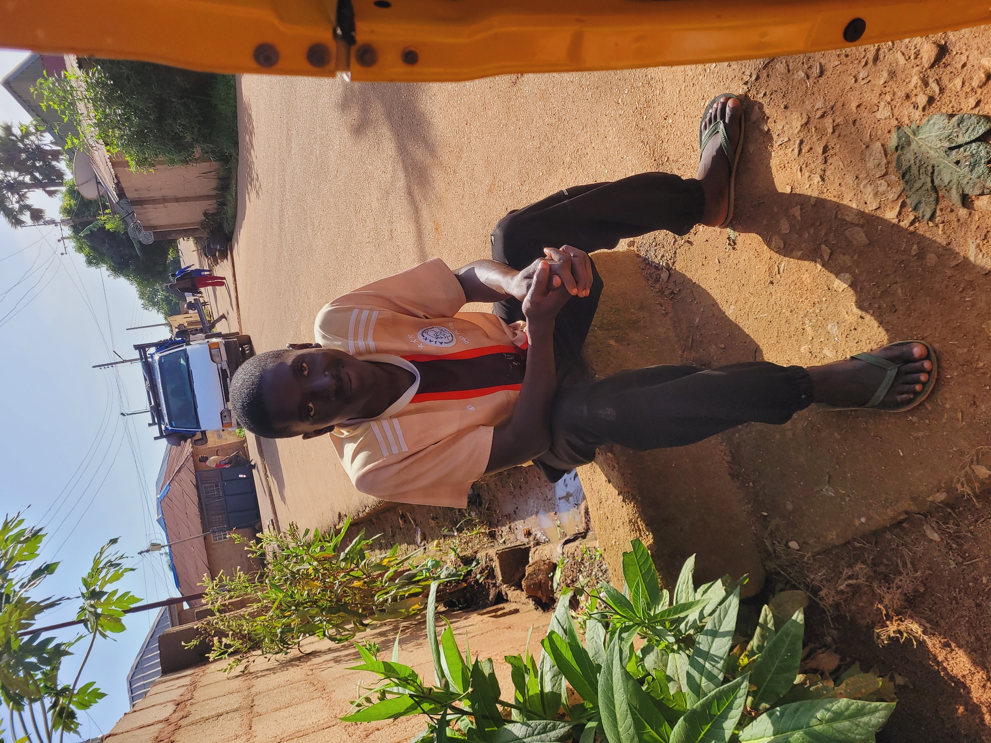 A young Black male is sitting on a concrete step in front of a drainage ditch, in what appears to be a rural area of Bukuru, Nigeria. He is wearing a light orange t-shirt with white stripes and dark pants. His hands are clasped in his lap, and he is wearing green flip-flops. The background shows a dirt road with a few buildings and a truck. The sun is shining, casting a shadow from the young man onto the road. To the right of the frame is a yellow vehicle, possibly a rickshaw.