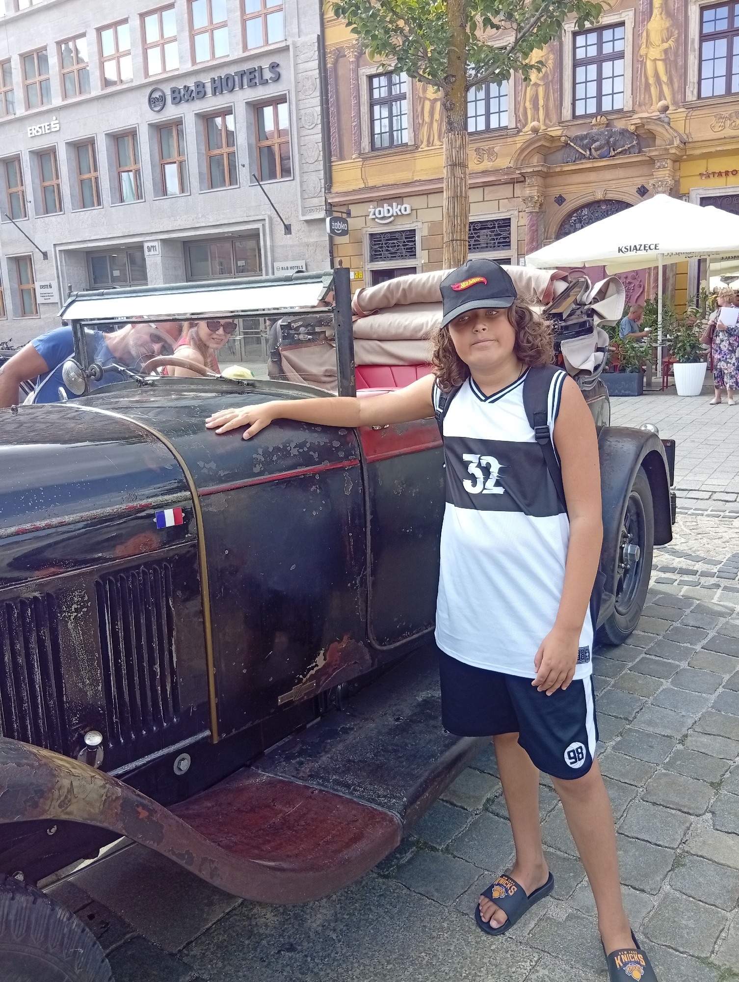 The image captures a young person standing next to a vintage black car in Wrocław, Poland. The young person, wearing a black baseball cap, a white and black jersey with the number "32" on it, and black shorts with the number "98" on the leg, stands with their right hand resting on the car. They are also wearing black slides with an orange basketball logo. In the background, two adults are visible inside the car. The environment suggests a street scene with cobblestone pavement and buildings, one of which displays "B&B HOTELS" signage. The car itself shows signs of age, with a French flag emblem on its front.