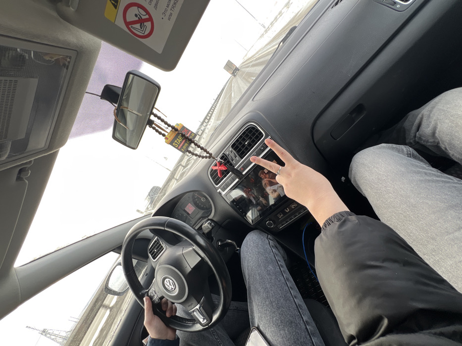 The scene depicts the interior of a Volkswagen vehicle, likely a Polo model, in the city of Khapry, Russia, viewed from the front passenger seat.

Two individuals are present: a driver and a front passenger. The driver's right hand, clothed in a blue sleeve, is gripping the black multi-function steering wheel on the left side of the frame. The driver's lower body, clad in light blue jeans, is visible. The front passenger's left hand, adorned with a ring on the fourth finger and emerging from a black jacket sleeve, is prominently featured in the foreground making a peace sign (V-sign). The passenger's legs, also in light blue jeans, are visible. A white smartphone rests on the passenger's left leg. A blue cable is visible near the passenger's right knee.

The dashboard is dark gray. The center console features an infotainment screen displaying what appears to be an image of two individuals. Air conditioning vents are located on either side of the screen. The instrument cluster is partially visible behind the steering wheel. A rearview mirror hangs from the windshield, from which a string of dark beads, a red cross-shaped ornament, and two rectangular air fresheners/tags are suspended. One tag is yellow with black text, prominently featuring "STAR" and a Russian phone number (e.g., "8 (919) 824-XX-XX"); the other is black with similar branding. The passenger-side sun visor is flipped up, revealing a warning label in Russian.

The vehicle is in motion on an outdoor road, likely a highway or major thoroughfare, with a visible metal guardrail on the right. The road surface appears clear but wet, with snow or ice accumulation on the shoulders and median. The sky is overcast and bright. In the distance on the left, a tall power line tower is visible. Other vehicles, including a truck, are discernible in the far distance, indicating traffic.