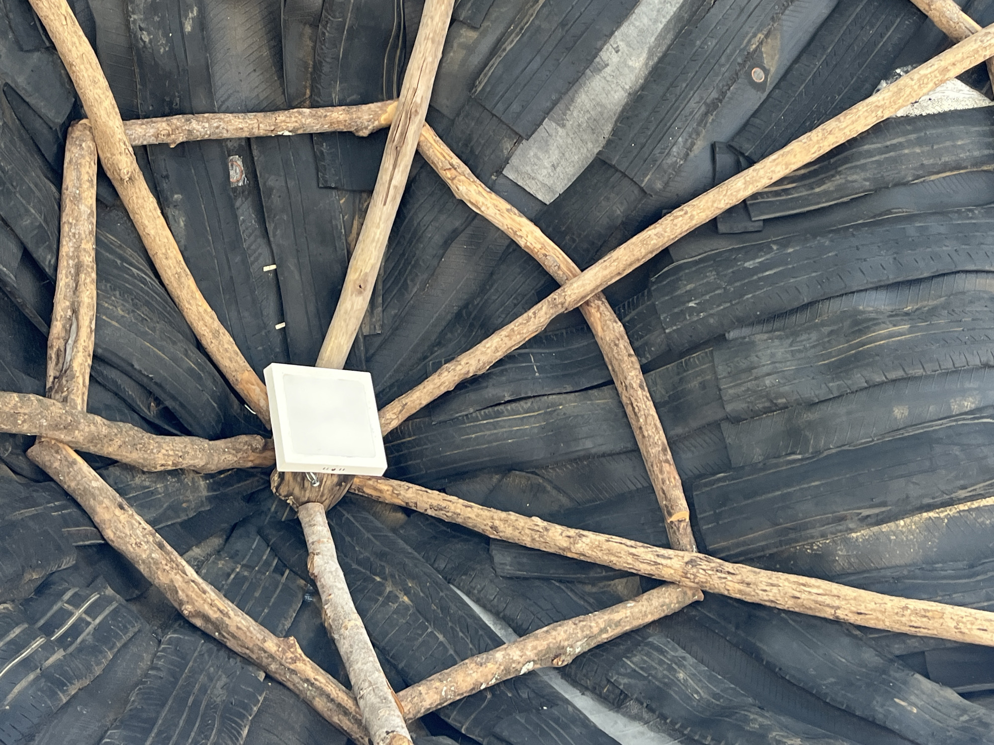This image shows a ceiling made of tires and wooden supports. The tires are black and arranged in an overlapping pattern, creating a textured surface. Wooden poles radiate outwards from the center of the ceiling, forming a geometric design. A white, square light fixture is mounted at the central point where the wooden poles converge. The photograph was taken in Juan Griego, Venezuela.