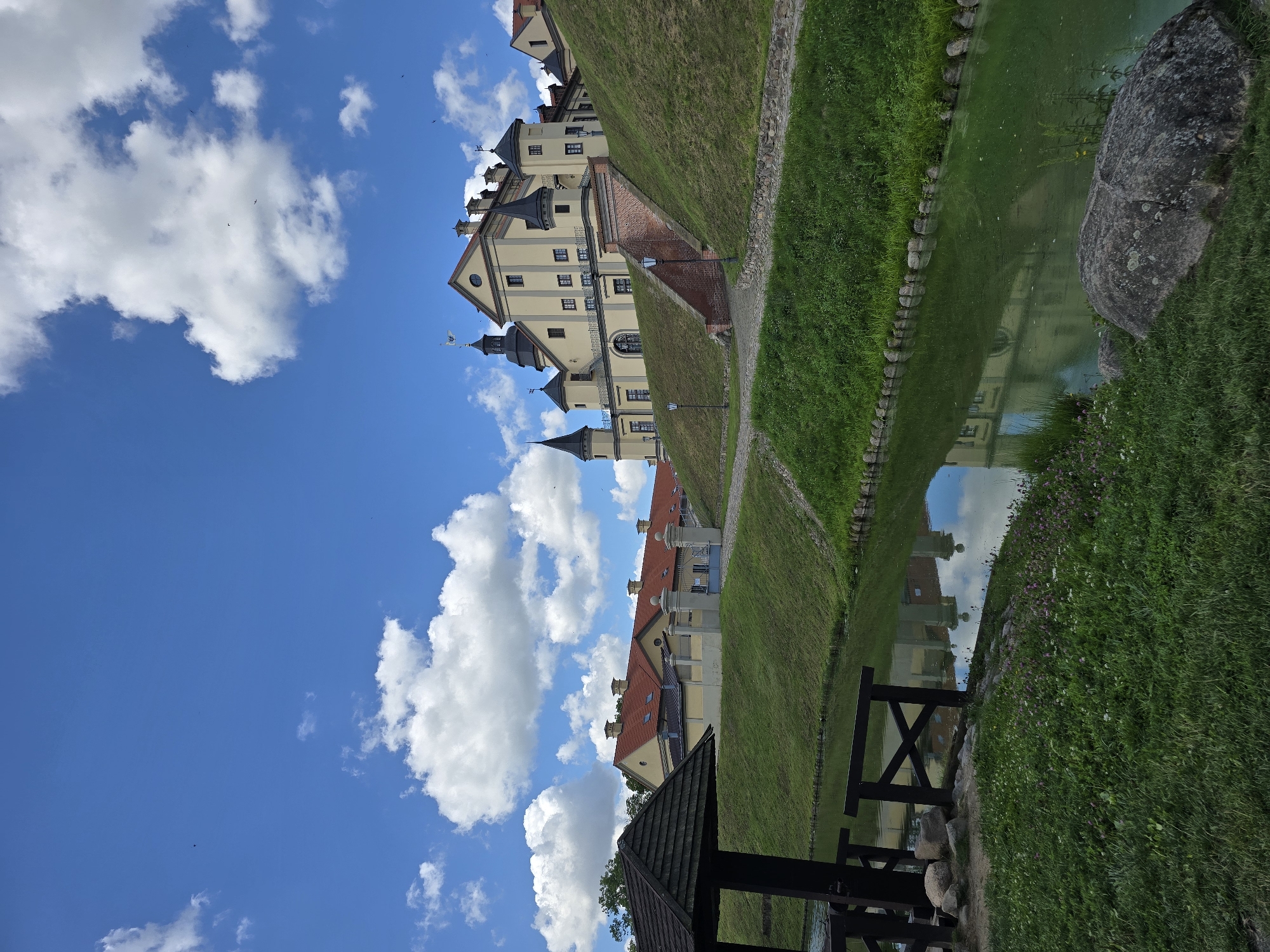 This is a vertical, outdoor, daytime, eye-level shot of Nesvizh Castle in Nesvizh, Belarus. The castle, built of pale yellow stone, sits atop a grassy mound. It has multiple towers with dark conical roofs, dormer windows, and a flag flying from one of the highest points. A red brick staircase ascends the mound to the castle entrance. A moat, with a green, murky surface reflecting the sky and the castle, surrounds the mound. In the foreground, to the left, is a dark wooden, open-sided structure with a peaked roof. A wooden railing is visible in the moat just below this structure. The grassy bank of the moat is dotted with small purple and white flowers. A large, grey boulder rests on the grass at the water's edge in the lower right corner. The sky is a vibrant blue with fluffy white clouds, and several small dark specks, possibly birds, are scattered across the sky.