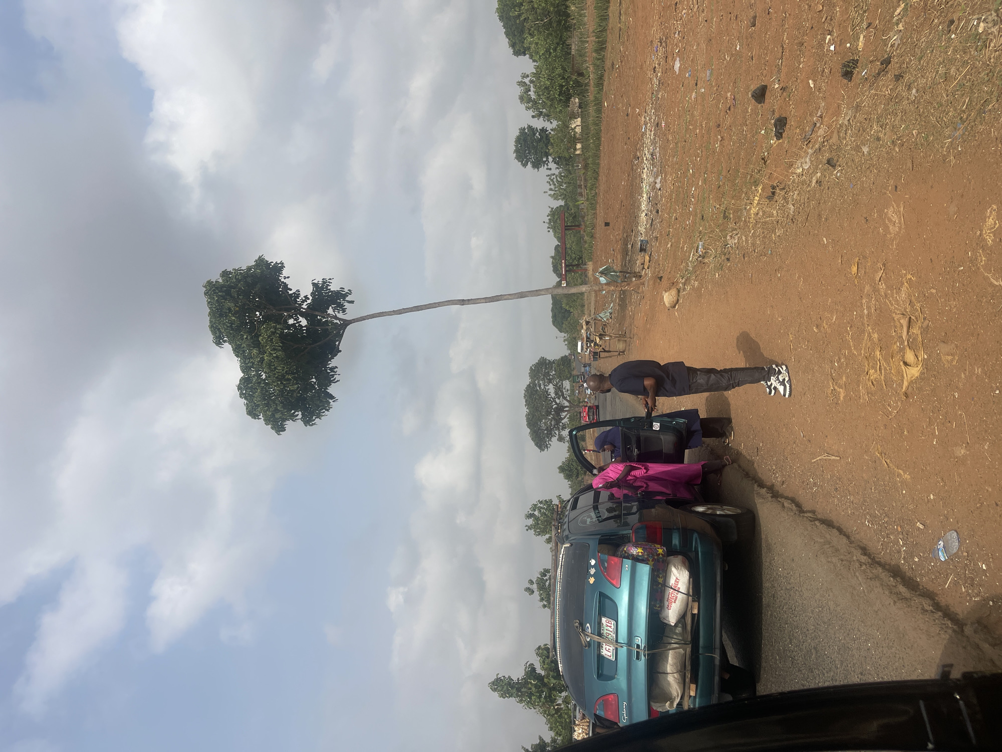 A blue minivan, license plate LFA 291 XB, is parked along the side of a narrow asphalt road in Karshi, Nigeria. Luggage is strapped to the roof of the minivan. A man in dark clothing stands near the driver's side, interacting with someone in the car. Another person in a pink garment is partially visible near the car. The road is bordered by dirt and sparse vegetation. In the background, several people are visible. A distinctive tall tree with foliage at its top stands out in the landscape. The sky is cloudy.