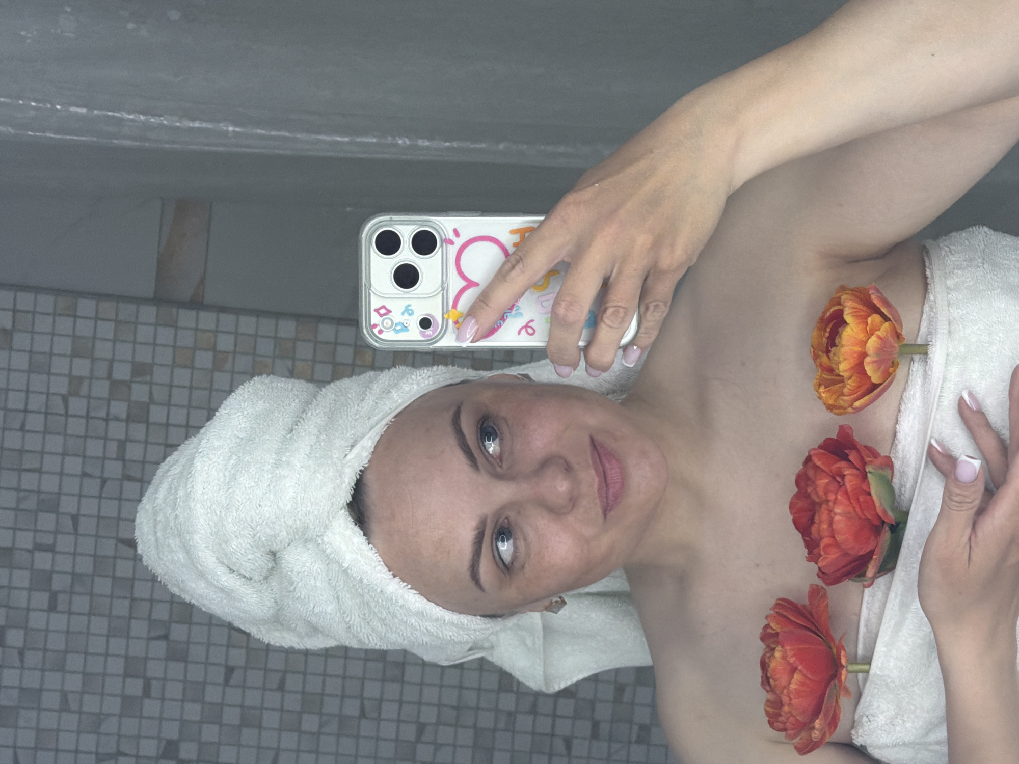 A woman with a towel wrapped around her head is taking a selfie in a bathroom. She holds a phone in her right hand, her fingers visible, showing a manicure. She is covered with a white towel, with three orange flowers placed on top. The background shows tiled walls. There is no specific information to indicate that this scene is located in Votkinsk, Russia.
