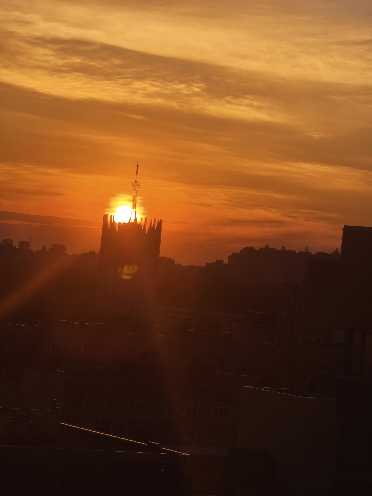 The image captures a sunset in Saint Petersburg, Russia, with the setting sun positioned directly behind the Saint Petersburg TV Tower, a tall broadcasting tower recognizable by its distinctive antenna. The tower's silhouette is prominent against the bright sun, with the sun appearing to be partially obscured by the tower's structure. The cityscape surrounding the tower is mostly in silhouette, suggesting a low-light environment and a long shot of buildings, with the rooftops of several buildings visible in the foreground. The sky is a gradient of warm colors, from orange to yellow, indicative of sunset conditions. The image does not depict any people, actions or interactions.