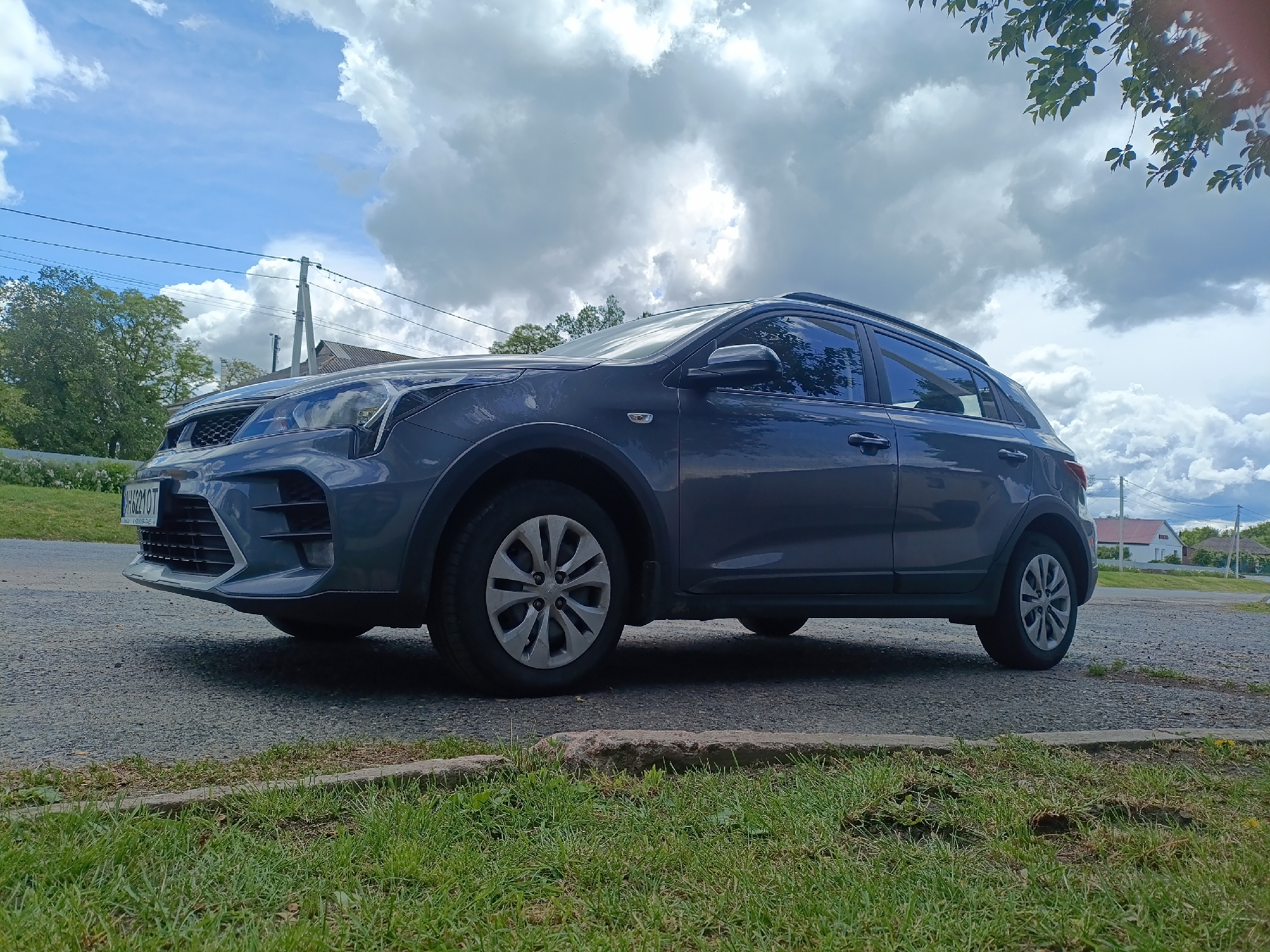 A dark grey Kia Rio X stands parked on a gravel road in Ustyvytsia, Ukraine. The car faces left, exhibiting its front end and side profile. Its license plate is visible. The car is surrounded by green grass on the foreground, with a curbstone separating the gravel road from the grass. The background includes houses with red roofs, trees, utility poles, and a cloudy sky. The perspective is a low-angle shot.