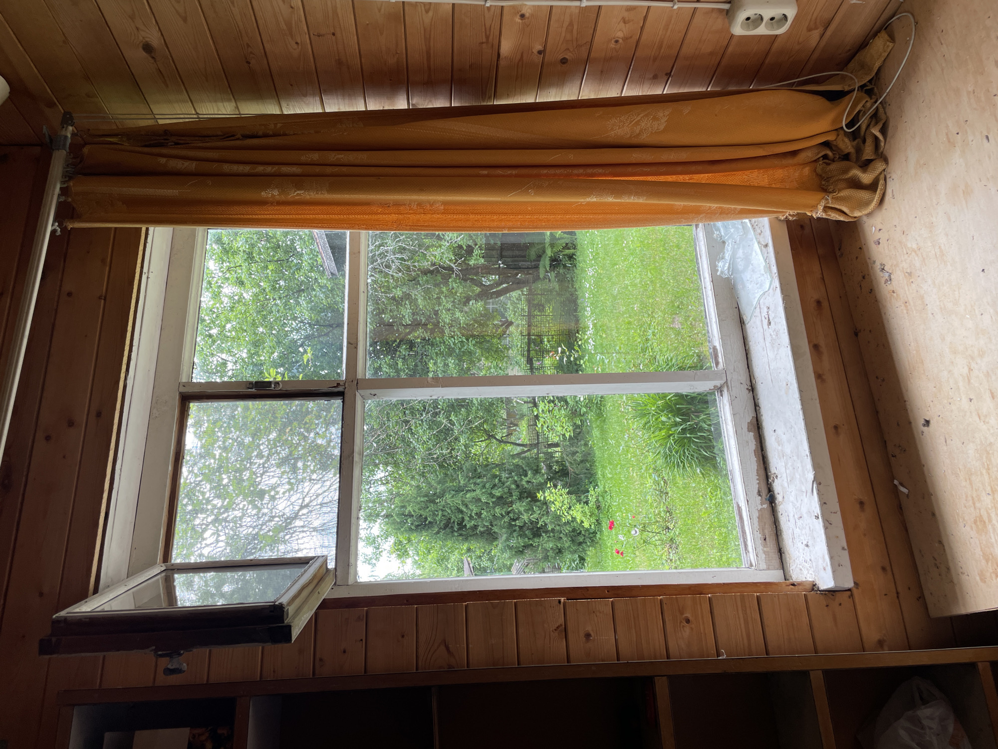 This media file shows an interior view of a room with wooden paneling on the walls and ceiling. The room appears to be a dwelling, possibly a cabin or a rustic home. The main focal point is a large window that looks out onto a green, grassy yard with trees. A worn, mustard-yellow curtain is partially drawn across the top of the window.
Visible objects include the window frame, which appears to be old and possibly in need of repair, with some peeling paint. A part of the window is open, revealing a screen. To the left of the window, a wooden shelf or cabinet is visible, with a glass-covered frame resting on it, slightly ajar. The ceiling has wooden planks and a visible electrical outlet with two sockets.
The scene is outdoors, with lush greenery and what looks like a fence or a structure in the background. There are also a few small red and white flowers visible in the grass. The time of day appears to be daytime, with natural light filtering through the window. The weather seems pleasant, as indicated by the vibrant green of the grass and leaves. No people are visible in the image. The overall impression is one of a simple, perhaps aged, rural dwelling. There is no visible text in the image.