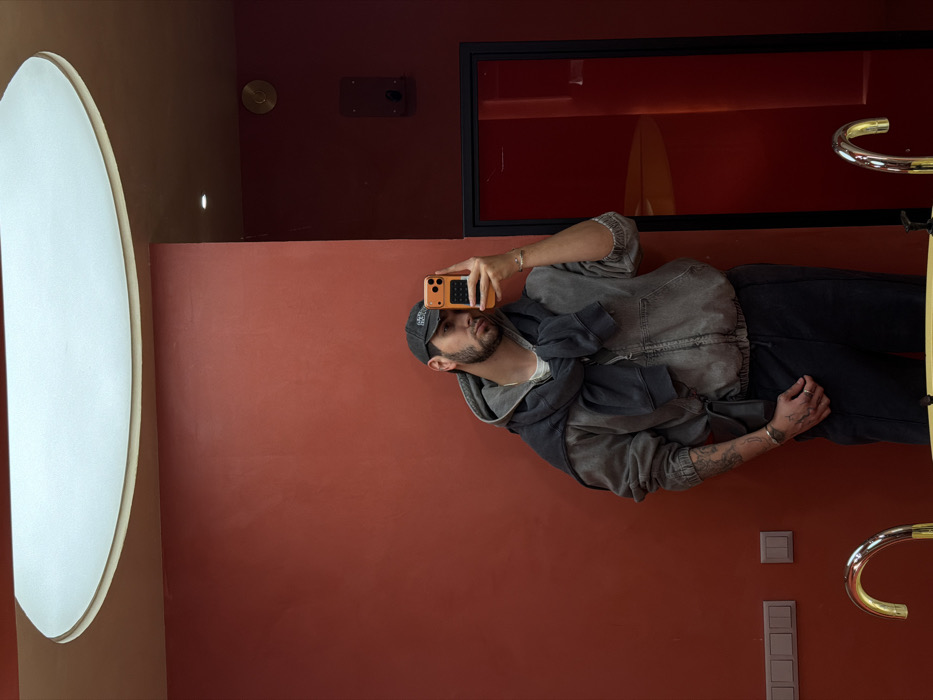 The image displays a male individual capturing a mirror selfie within an indoor establishment in Yerevan, Armenia, likely a modern restroom or vanity area. The specific name or address of the location is not identifiable from the visual information provided.
The subject is a man with a beard and dark hair, wearing a dark baseball cap with a white emblem, a light-colored t-shirt, a grey full-zip hooded jacket, and dark trousers. A darker grey sweatshirt is casually tied around his neck and shoulders. Visible tattoos adorn his left forearm, and he wears a silver-colored bracelet on his left wrist and a ring on his left hand. He holds an orange-cased smartphone in his raised right hand, directing its lens towards his reflection in the mirror.
The surrounding environment features smooth, reddish-brown painted walls and a lighter, tan-colored ceiling. A large, elliptical recessed light fixture is prominently centered on the ceiling. On the lower left wall, a white triple-gang light switch panel is installed. To the upper right, a dark rectangular panel with a small black component and a small, circular brass fixture above it are mounted. The far right side of the frame shows a dark-framed opening or recess, revealing a darker, red-hued interior space with a reflection of a light source. In the foreground at the bottom, two golden gooseneck faucets and portions of a yellow sink basin are visible, indicating a wash area.