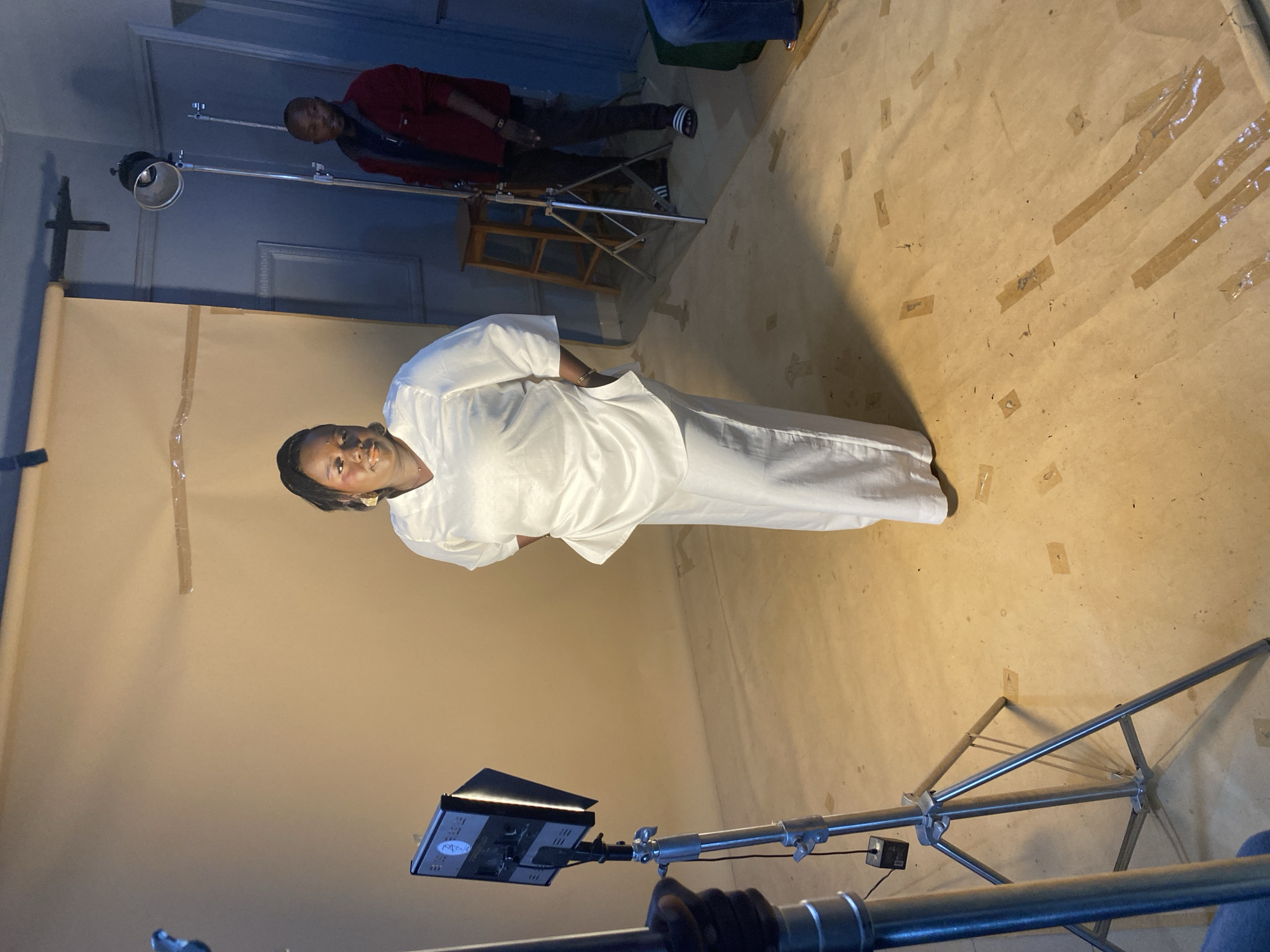 In a studio setting in Bukuru, Nigeria, a woman wearing a white outfit stands facing the camera. She has dark hair and is looking directly at the viewer. To her left, a man in a red jacket and dark pants stands behind a lighting stand. Another person, whose legs are visible in the upper right corner, is also present. The floor is covered with a light-colored backdrop that is taped down in several places. In the foreground, a camera light on a stand is positioned towards the woman. The overall scene appears to be a professional photo or video shoot, with the lighting equipment and backdrop indicating a controlled environment.