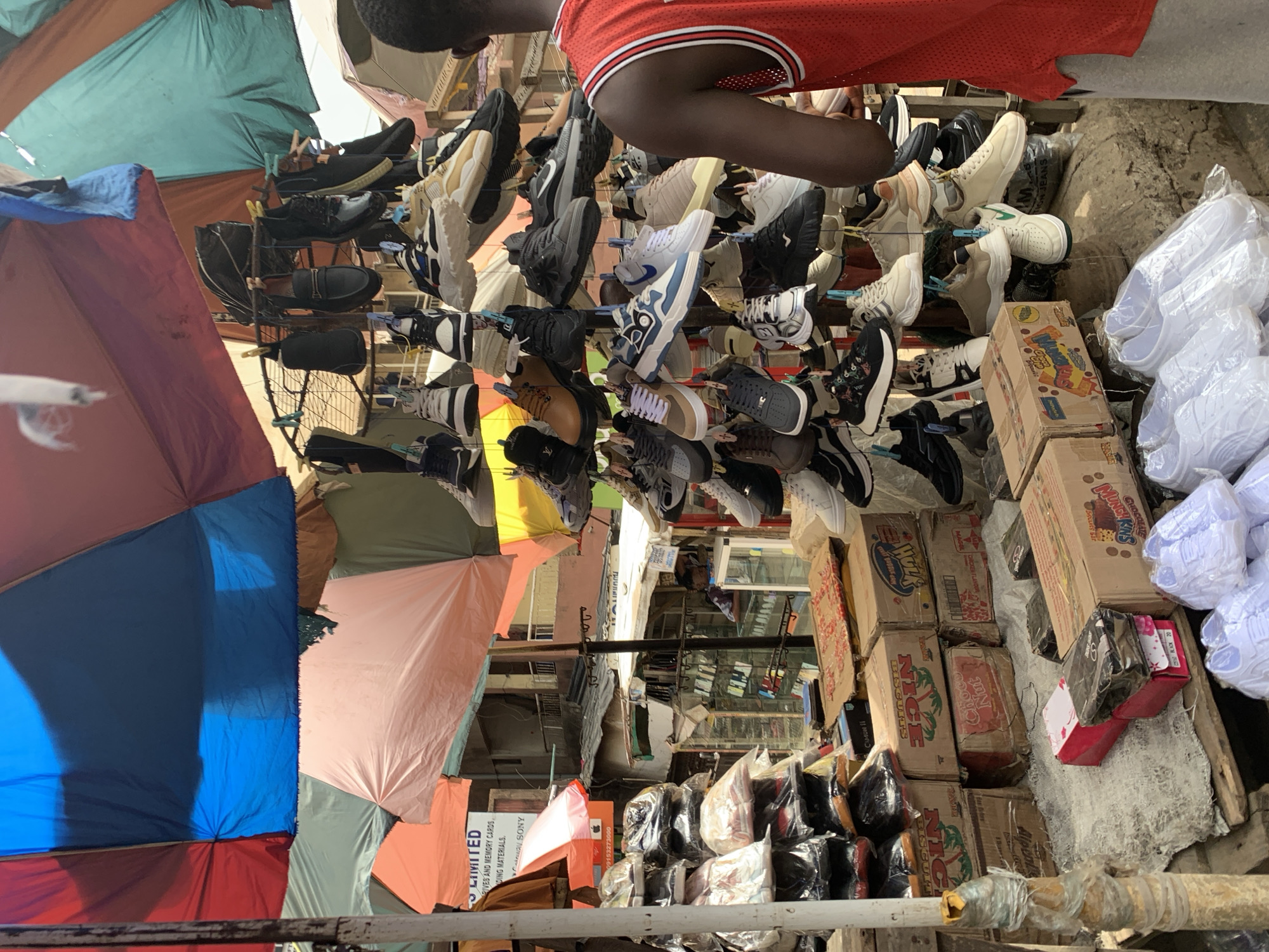 The image depicts an outdoor market scene, likely in Ikorodu, Nigeria. Hanging rows of assorted sneakers and shoes are suspended from overhead wires and structures, using clothespins. A person wearing a red and white basketball jersey partially obscures the view, suggesting they are browsing or working at the stall. The vendor's hand is shown reaching for a shoe. Below the hanging shoes, various cardboard boxes, some labeled "Nice Biscuits," and other merchandise are piled on a wooden platform. Several colorful umbrellas provide shade to the vendors and shoppers, creating a patchwork canopy above. A building is visible in the background, giving clues to the urban location. The general atmosphere is busy and bustling.