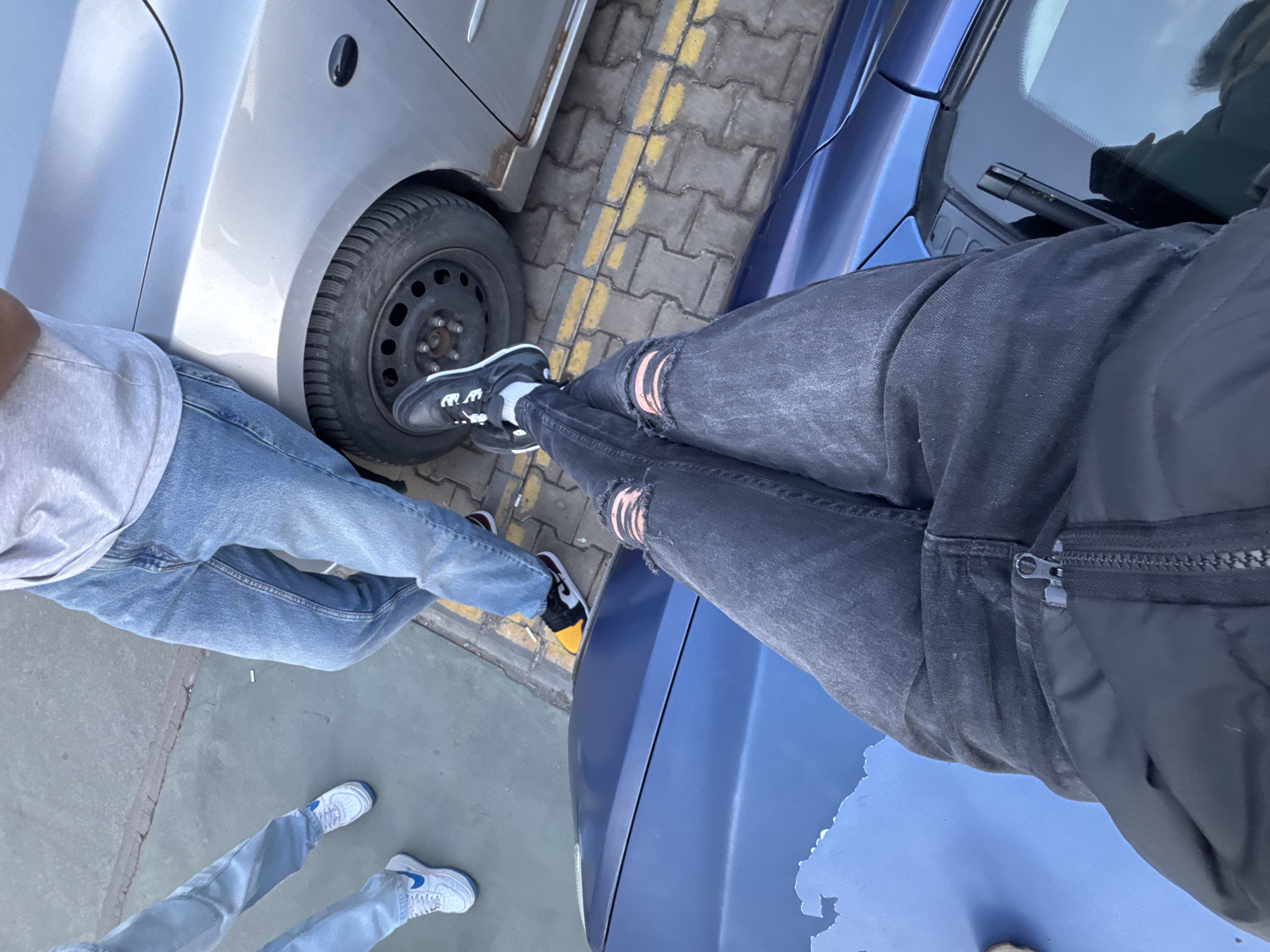 This high-angle image captures the lower bodies of at least three individuals in a casual, outdoor setting in Sofia, Bulgaria. The perspective looks down at their legs, feet, and the surrounding environment, primarily featuring two cars and a paved street.
In the foreground on the left, a person wears light blue jeans and white sneakers with blue accents, standing on light grey concrete. Another individual, partially visible on the mid-left, is wearing light blue jeans and a grey t-shirt, with their left foot positioned near the front wheel of a silver hatchback car. Their shoes are black, yellow, and red. To the right in the foreground, a third person is seen wearing dark grey ripped jeans, a black jacket, and black and white sneakers, with their right foot resting on the hood of a bright blue car.
The silver car features a black steel wheel fitted with a winter tire. The blue car, which is closer to the viewer, exhibits noticeable wear with areas of peeling paint on its hood. The ground beneath them is a mix of smooth concrete and grey interlocking pavers, with a distinct double solid yellow line running across the pavers, typically indicating parking restrictions. A white cigarette butt is visible on the concrete.
The scene takes place during bright daylight, suggesting sunny weather. The overall impression is a casual stop or gathering in an urban or suburban street environment. No explicit text is clearly visible on clothing or objects.