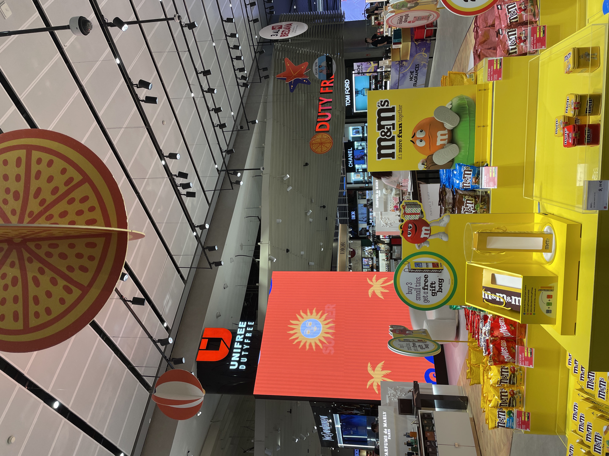 The image captures a brightly lit interior, likely a duty-free shop, with prominent displays of candy and luxury goods. A large, vibrant orange and yellow circular decoration hangs from the ceiling, reminiscent of a sliced citrus fruit. Below this, a large red digital screen displays a stylized sun with the word "SUMMER" beneath it, flanked by palm trees.
To the left, a red and white striped spherical decoration hangs in the air. Above this, the prominent red "U" logo of "UNIFREE DUTY FREE" is visible on a black pillar. Adjacent to the digital screen, a display for "YVES SAINT LAURENT" is partially visible. Further to the right, signs for "DIOR," "CHANEL," and "TOM FORD" indicate a luxury retail environment.
The foreground is dominated by a yellow display featuring M&M's candy. Several bags of M&M's and Maltesers are arranged on shelves, with promotional signage indicating "BUY 3 small tins, get a free gift bag." A large M&M's character stands next to the display. Below, a yellow counter showcases small M&M's tins. The overall impression is a commercial setting designed to attract shoppers with bright colors and enticing promotions. The location is identified as Arnavutköy, Turkey, suggesting this is an airport duty-free or a similar retail establishment within that region.