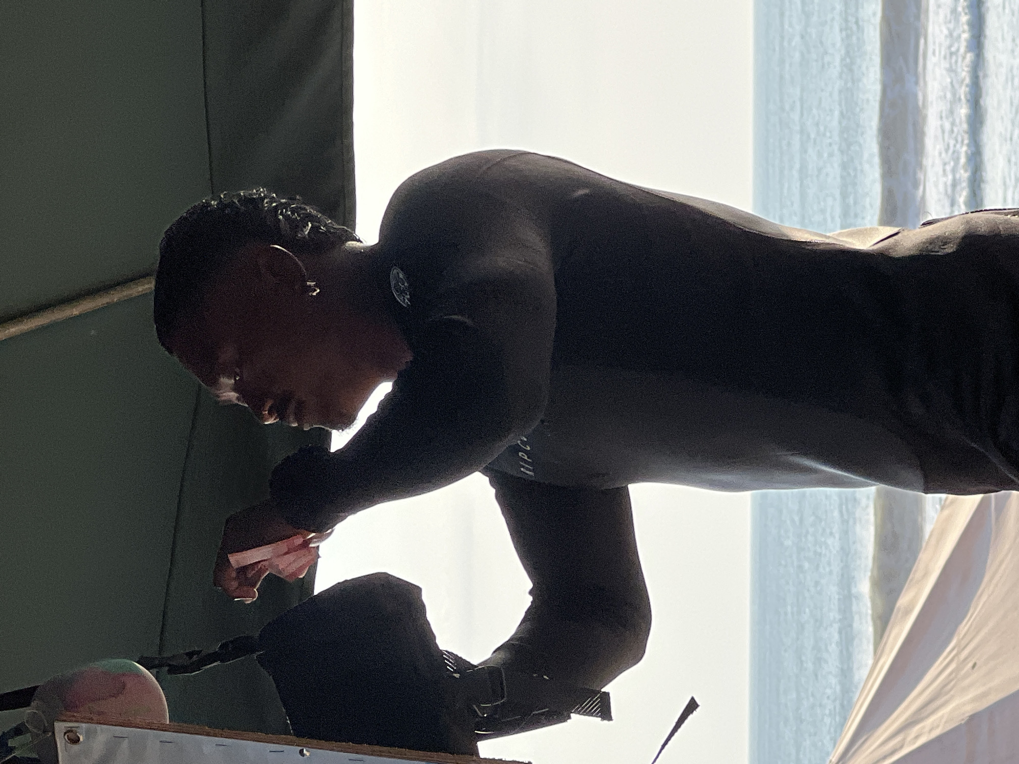 The image shows a Black man wearing a black wetsuit, standing outdoors with the ocean and beach in the background. He is looking upwards and to his right, holding a small, pink item in his hands, possibly a piece of paper or fabric. His hair is styled in short, textured braids. He is also wearing a silver hoop earring in his left ear. The man appears to be on a beach or near the water, possibly preparing for or finishing a water sport. A white tarp or shelter is visible in the foreground, and a wooden structure with a metallic object attached to it is on the left side of the frame. The lighting suggests it is daytime, with the sun casting some shadows and illuminating the scene. The ocean has moderate waves. The overall impression is of a casual, outdoor activity near the sea. Kuta, Indonesia is a popular beach destination known for its surfing and lively atmosphere.