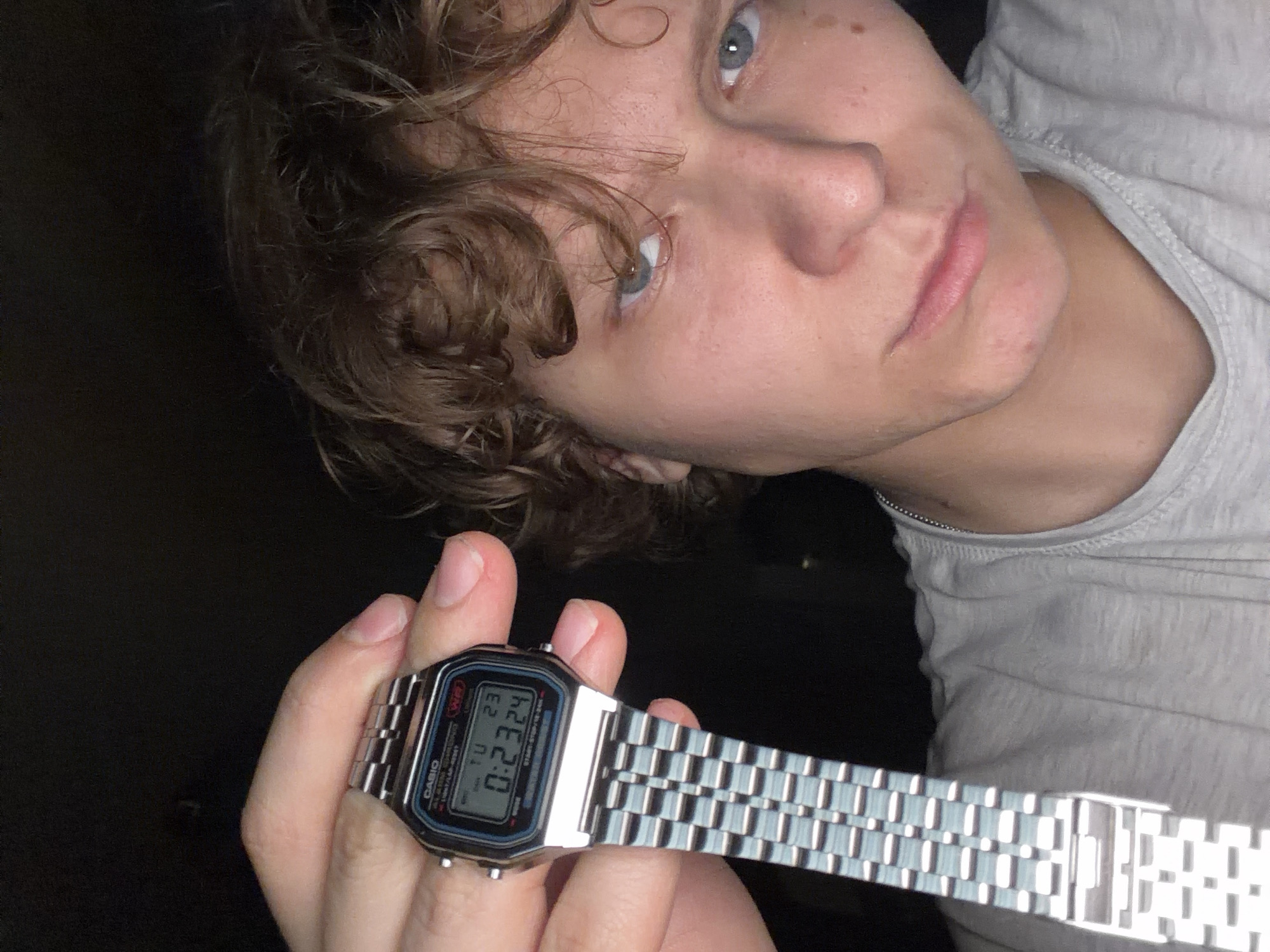 The image shows a close-up of a young person holding up a silver Casio digital watch to the camera. The watch face displays the time as 0:23:24, with "TU 23" also visible, possibly indicating Tuesday the 23rd. The person has light skin, curly brown hair, and blue eyes. They are wearing a grey t-shirt and a thin chain necklace. The background is dark and indistinct. There are no discernible elements in the image that would suggest a location in Chistopol, Russia.