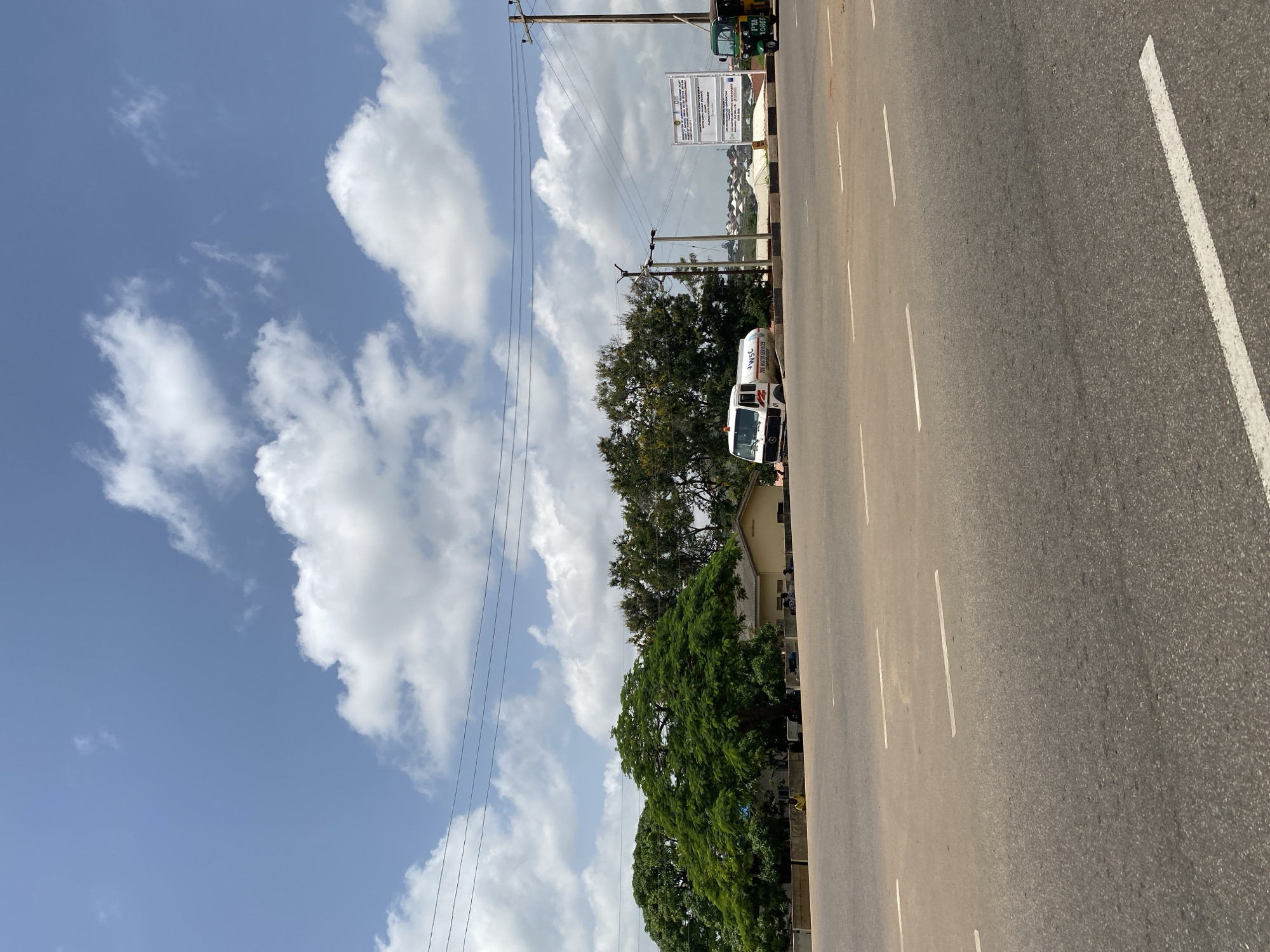 The image captures a bright, sunny day in Jos, Nigeria, with a clear blue sky dotted with fluffy white clouds. In the foreground, a wide, paved road with white lane markings dominates the lower half of the frame. On the left side of the road, a white tanker truck labeled "JOS WATER SERVICES CORPORATION" is parked. Behind the truck, a large, lush green tree with a wide canopy stands prominently. Further back, a low-slung beige building is visible, with a few people seated or standing near its base. A tall utility pole with wires stretches across the upper right section of the image. To the right of the tanker truck and in the background, a large white billboard with text and graphics is erected, and a green and yellow auto-rickshaw is parked next to it. The overall scene suggests an urban or semi-urban environment in Jos, likely near a water service facility.