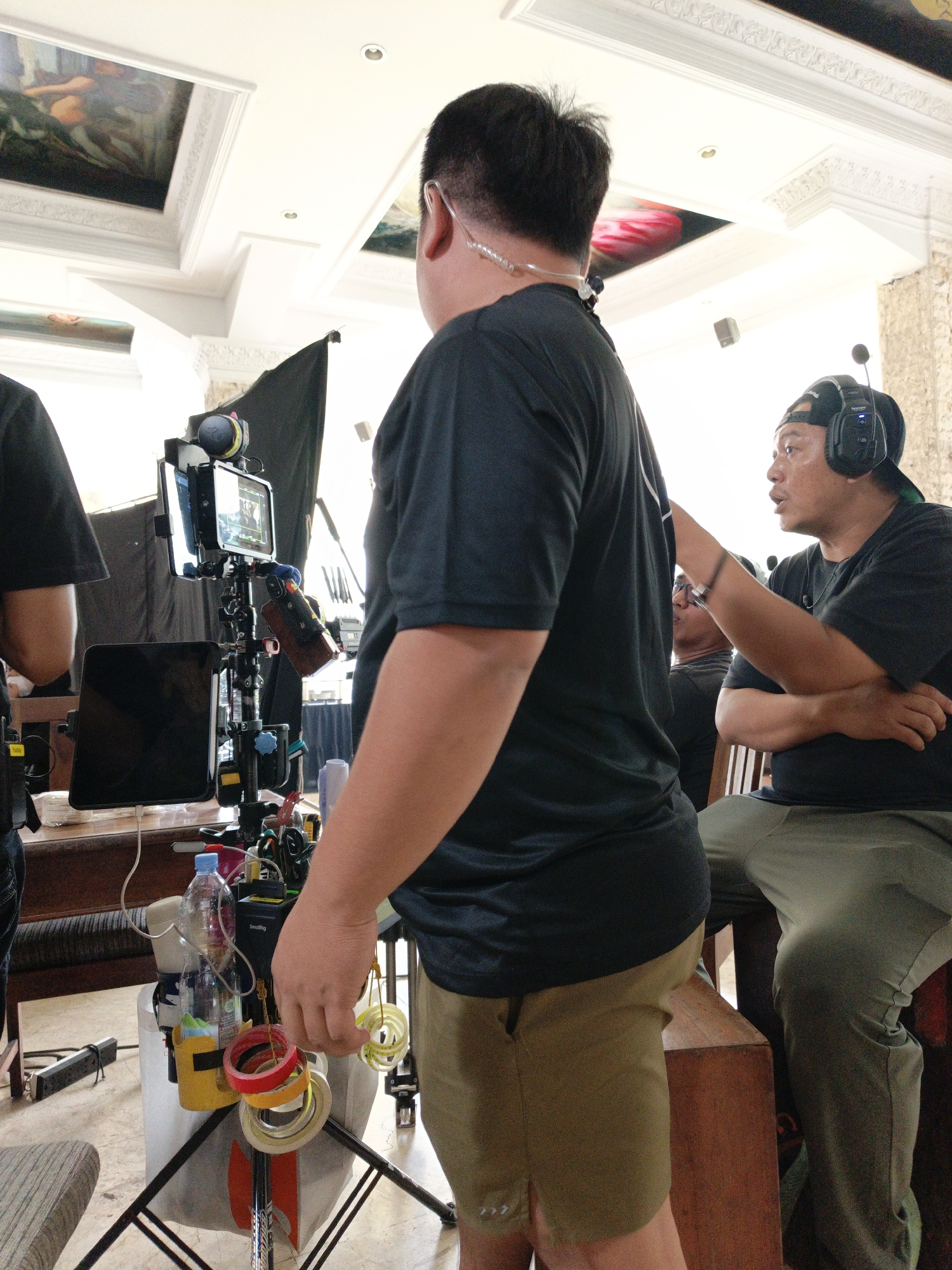 The image depicts a film set in Cibogo, Indonesia, likely an indoor location given the presence of elaborate ceiling decorations with paintings. In the foreground, a man wearing a black t-shirt and khaki shorts stands with his back to the camera, facing a complex film rig. This rig supports various electronic devices, including monitors and cameras, along with cables and rolls of tape. To his right, another man, wearing a black t-shirt, a baseball cap, and headphones, is seated and speaking, gesturing with his right hand towards the rig or the first man. A third person, partially visible behind the seated man, appears to be wearing glasses and a dark t-shirt. The environment suggests a professional production setting, with attention to equipment and lighting. The ceiling features ornate plasterwork and what appear to be large, framed paintings. The floor is tiled or made of stone. The overall scene captures a moment of active work on a film set.