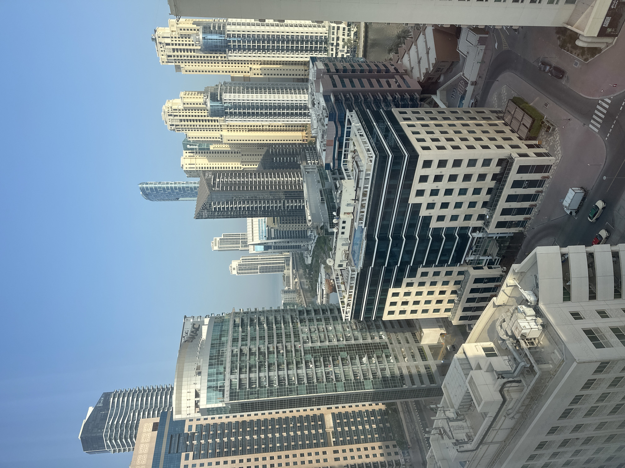 The image shows a high-angle view of a dense urban landscape with numerous skyscrapers in Dubai, United Arab Emirates. The architecture is predominantly modern, featuring glass facades, balconies, and sharp geometric shapes. Buildings are of varying heights and designs, with some having distinct curved or angular profiles. In the foreground, a large white building with visible HVAC units on its rooftop is prominent. Below it, a multi-story building with a dark glass facade and white accents is visible. The street level shows roads, sidewalks, and a few parked cars and trucks. Further into the background, a body of water, likely a canal or a creek, is visible with a boat docked. More skyscrapers of various designs and colors stretch into the distance, under a clear blue sky. The scene conveys a sense of a highly developed and affluent city.