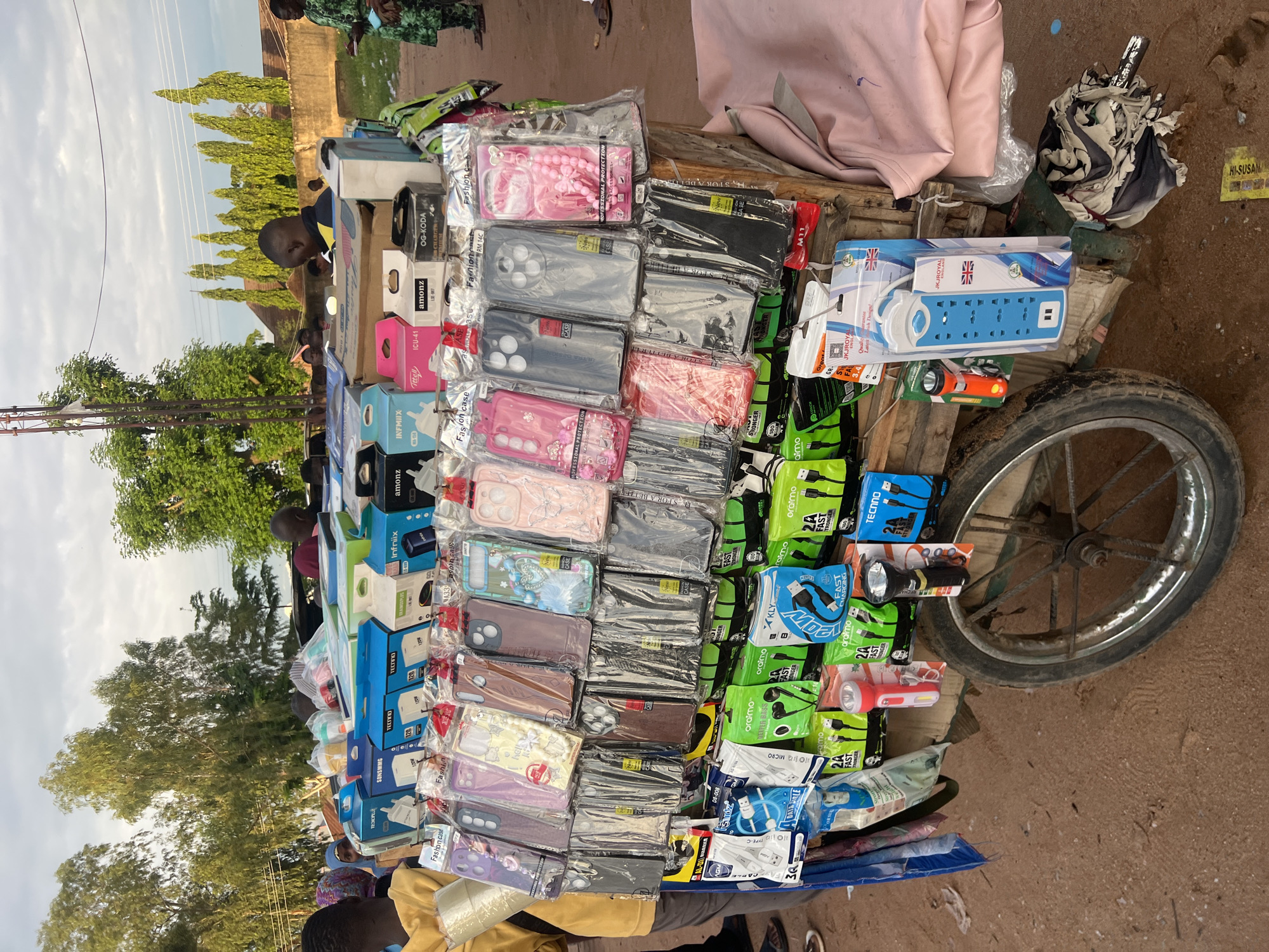 This image captures a street vendor's mobile stall in Jalingo, Nigeria, laden with electronic accessories. The central focus is a wooden two-wheeled cart, with one spoked wheel visible on the lower right, displaying various products.
The top tier of the cart features an array of boxed electronic items, likely chargers or power banks, from brands such as Tecno (notably 120W models), Sonsmmg, Fttell (ICU-41), Amonz (LU-987), OG-KODA, and Infinix (AR-480).
Below these boxes, numerous smartphone cases are hung, individually packaged in clear plastic sleeves. These "Fashion cases" display a variety of designs and colors, including purple, pink, brown, and black, with some featuring decorative elements like butterflies, flowers, or beaded strings.
The bottom section of the cart showcases smaller packaged accessories. These include various charging cables and earphones from brands like Oraimo (marketed as "Fast Stronger"), KLY ("Fast Charging"), and Tecno. Additionally, two flashlights (one red and white, one black) are visible, along with a blue multi-socket power strip with USB ports, branded "JKROYAL" and featuring a UK flag motif.
Several individuals are present in the scene. On the left, a person in a yellow top stands near the cart. In the background, other people are partially visible, including one in a patterned green outfit on the far right. The ground is unpaved, consisting of reddish-brown dirt or sand. The background features several trees, including tall, slender trees on the right and denser foliage on the left. A partial view of a building and an overhead utility pole/line structure are also discernible against a partly cloudy sky. The lighting suggests daylight, possibly late afternoon.