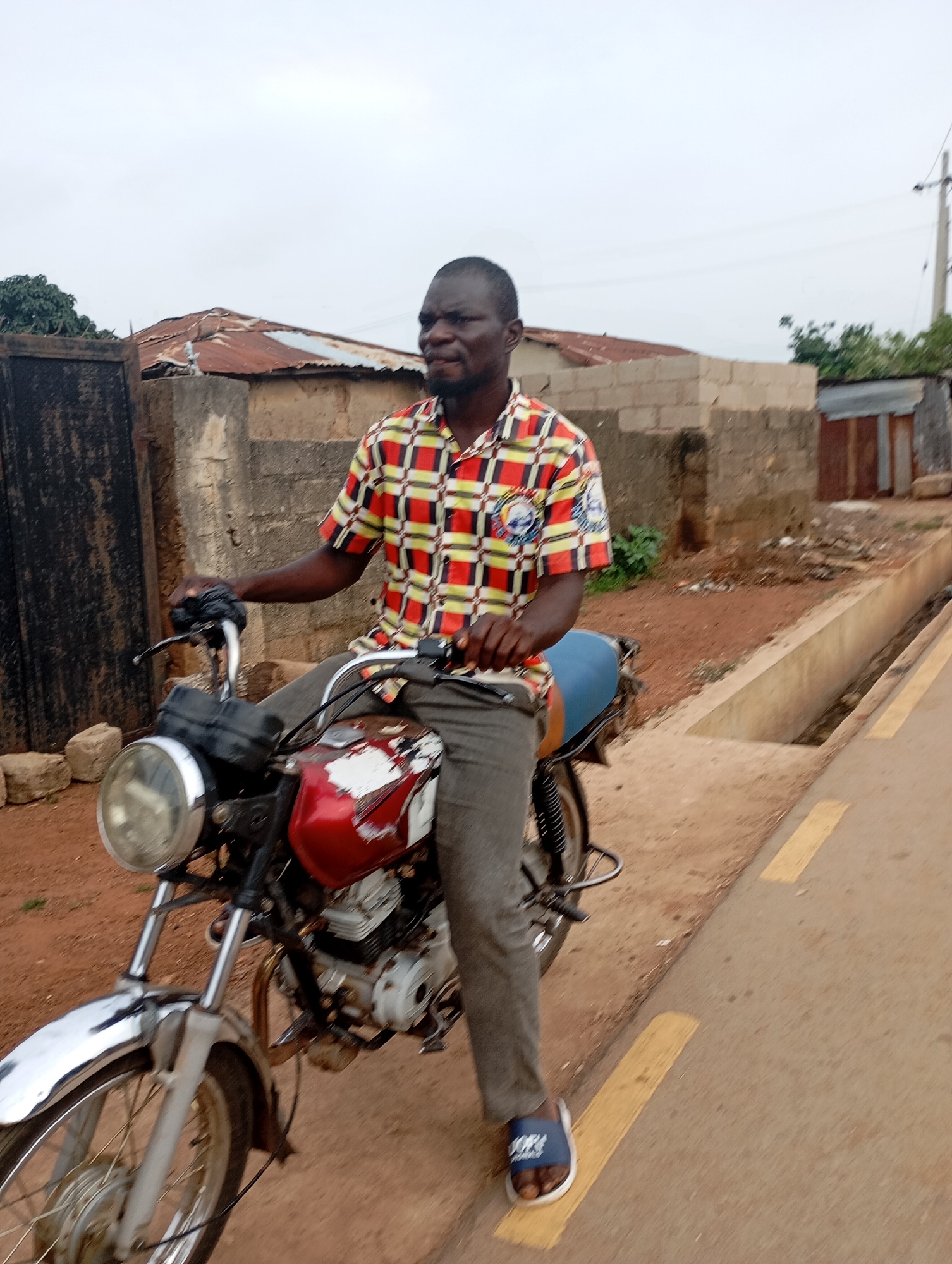 A Black man, wearing a red, yellow, and white checkered shirt and grey pants, is sitting on a red and white motorcycle. He is looking to the right and has his left hand on the handlebars. The motorcycle has a round headlight and a blue seat. The man is wearing blue sandals. He is on a dirt road, with a concrete ditch and yellow painted lines to his right. In the background, there are buildings with corrugated metal roofs and a brick wall. The sky is overcast. This scene is located in Shendam, Nigeria.