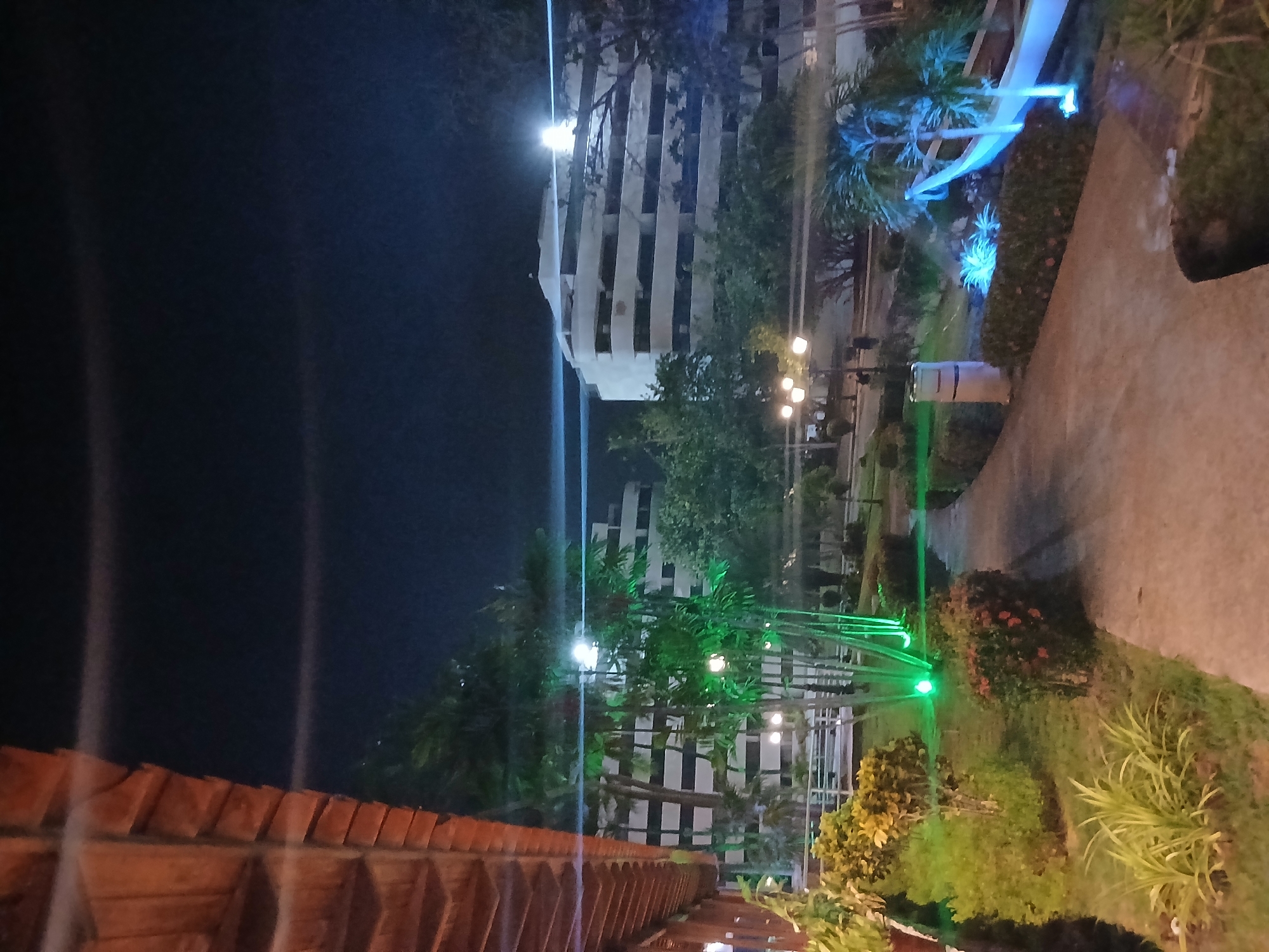 This media file presents a vibrant outdoor scene captured at night, likely within a resort or hotel complex in Playa El Agua, Venezuela. The image is oriented vertically but depicts a horizontal landscape.
In the background, tall, multi-story buildings with numerous windows or balconies are visible, illuminated from within and possibly by exterior lighting, highlighting their white or light-colored facades. The dark night sky fills the upper portion of the frame.
The foreground and mid-ground are characterized by a well-maintained landscaped garden. Lush vegetation, including tropical trees, palm trees, and various bushes and plants, fills the space. A light-colored paved pathway or sidewalk winds through the garden on the right side of the frame.
The scene is dramatically lit by a combination of colorful and white artificial lights. Bright blue lights illuminate several palm trees and sections of the greenery, while vivid green lights highlight other trees and a metal structure, possibly an archway or trellis, creating a striking contrast. Warm yellow lights from garden lamps softly illuminate portions of the pathway and imply the presence of outdoor seating areas, though specific furniture is indistinct. Bright white light sources, possibly from streetlights or reflections, create prominent vertical streaks across the image.
In the immediate foreground, a reddish-brown tiled roof structure, possibly part of a building or a covered walkway, is partially visible on the left. A light-colored cylindrical object, potentially a trash bin or planter, stands beside the pathway in the mid-ground.
There are no people visible in the image. The setting suggests a peaceful evening ambiance, designed for relaxation and aesthetic appeal in a tropical or coastal environment. No visible text is present.